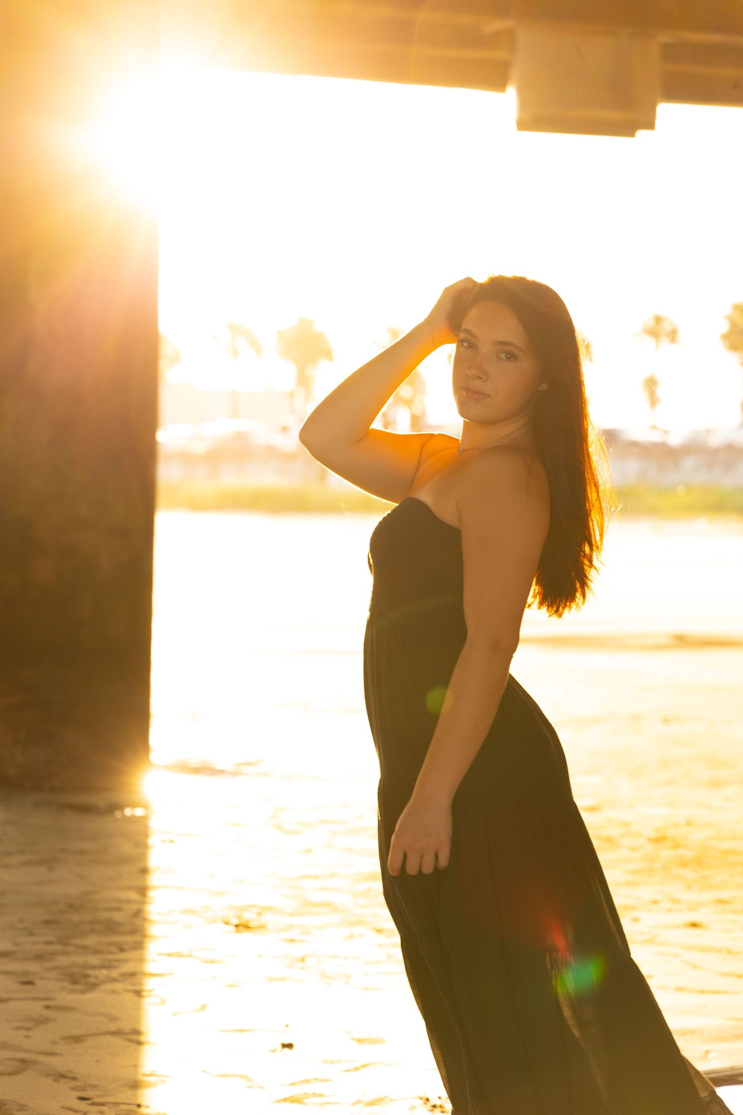 Woman in black dress poses gracefully under bridge at golden hour sunset with warm lens flare and palm trees in background.