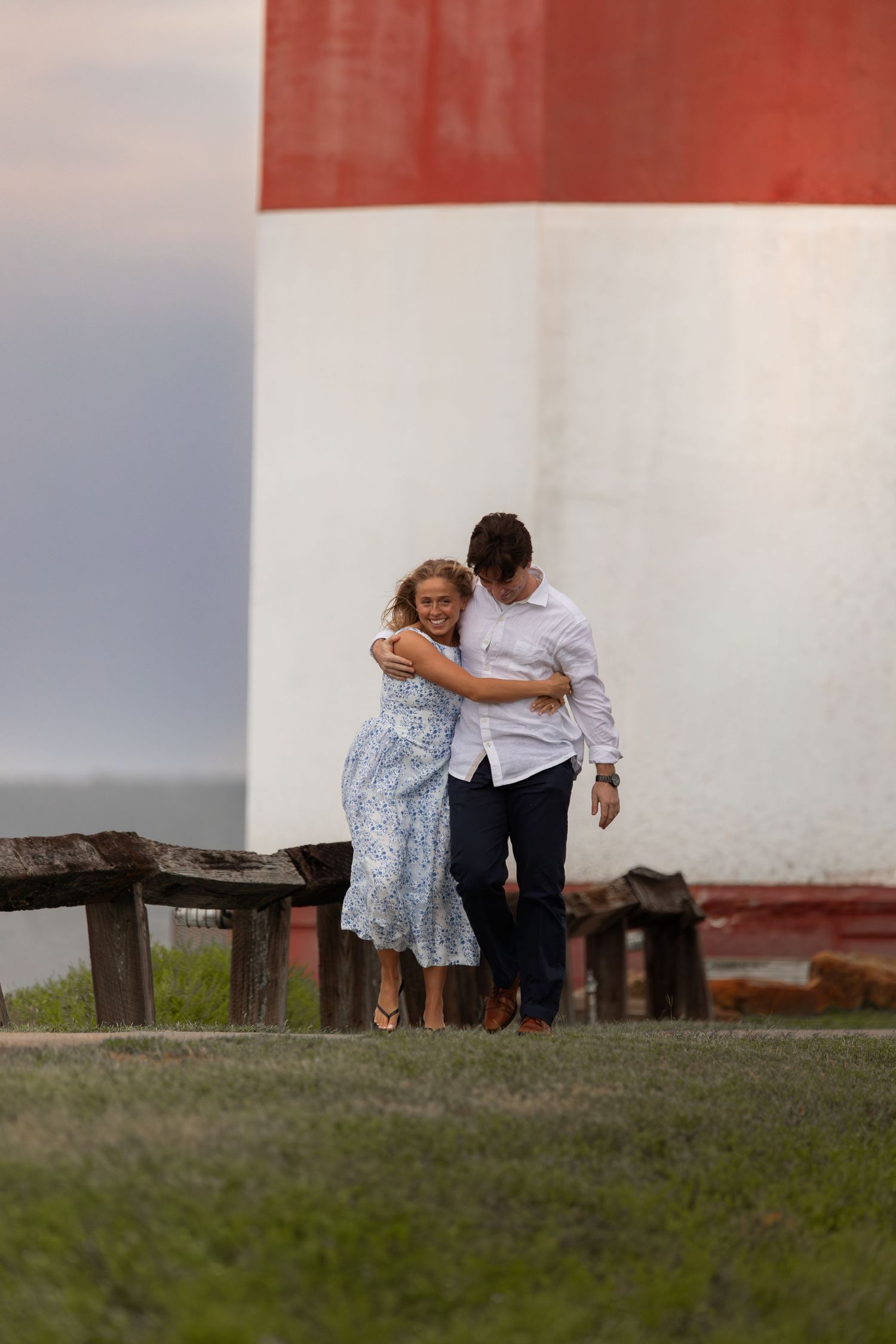 Couple embracing and walking together near a red and white lighthouse on a grassy coastal area.