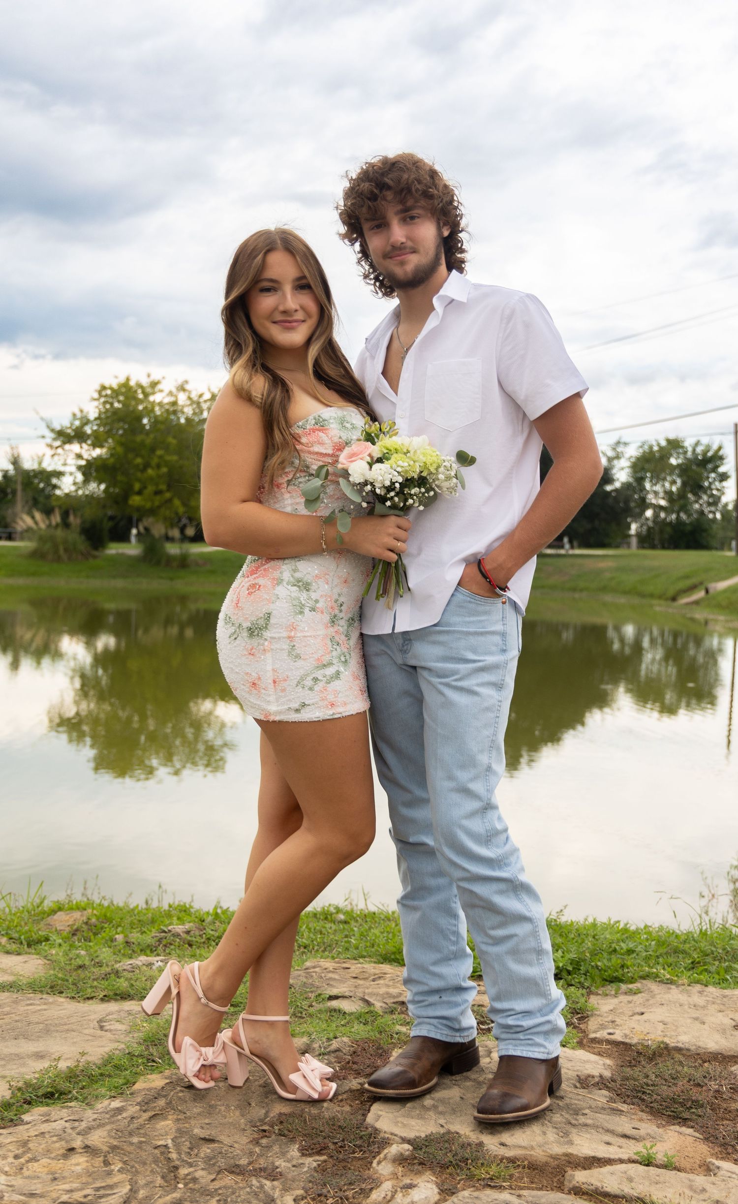 Young couple posing by a pond, woman in floral mini dress holding bouquet, man in white shirt and jeans.