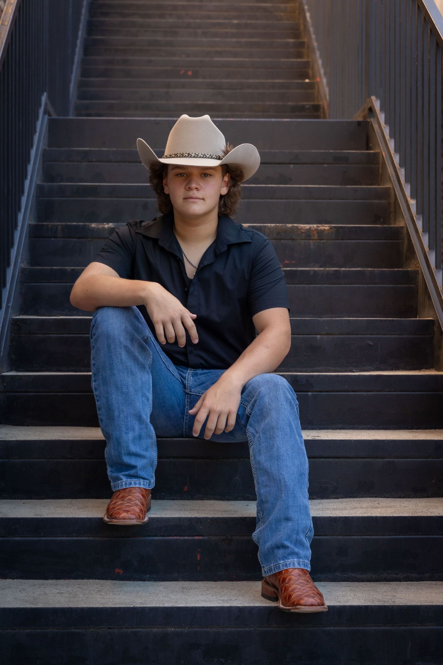 Young man in cowboy hat, black shirt, jeans, and boots sitting on dark metal stairs.