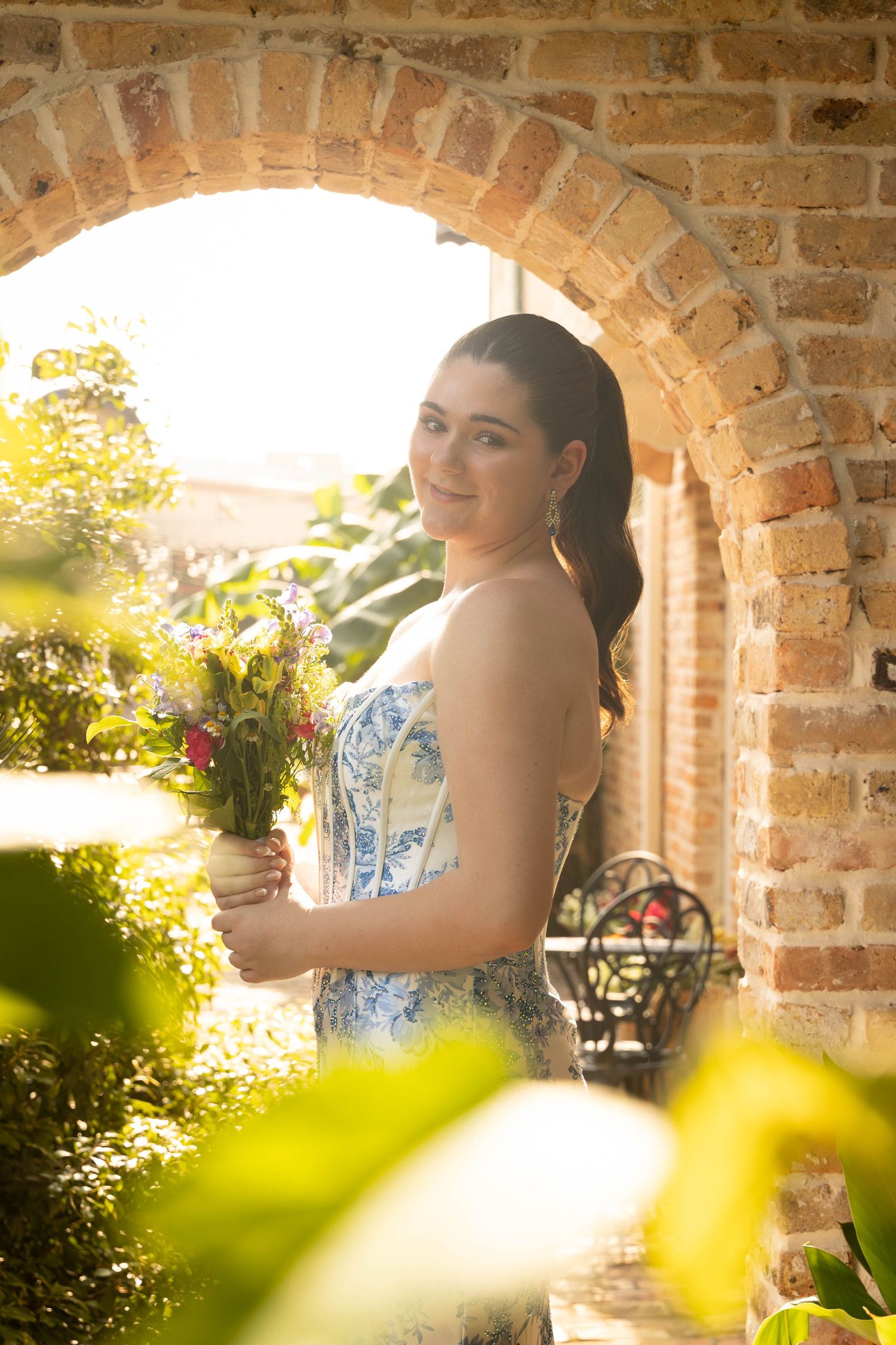 Young woman in floral dress holding wildflowers under a sunlit brick archway surrounded by lush greenery.