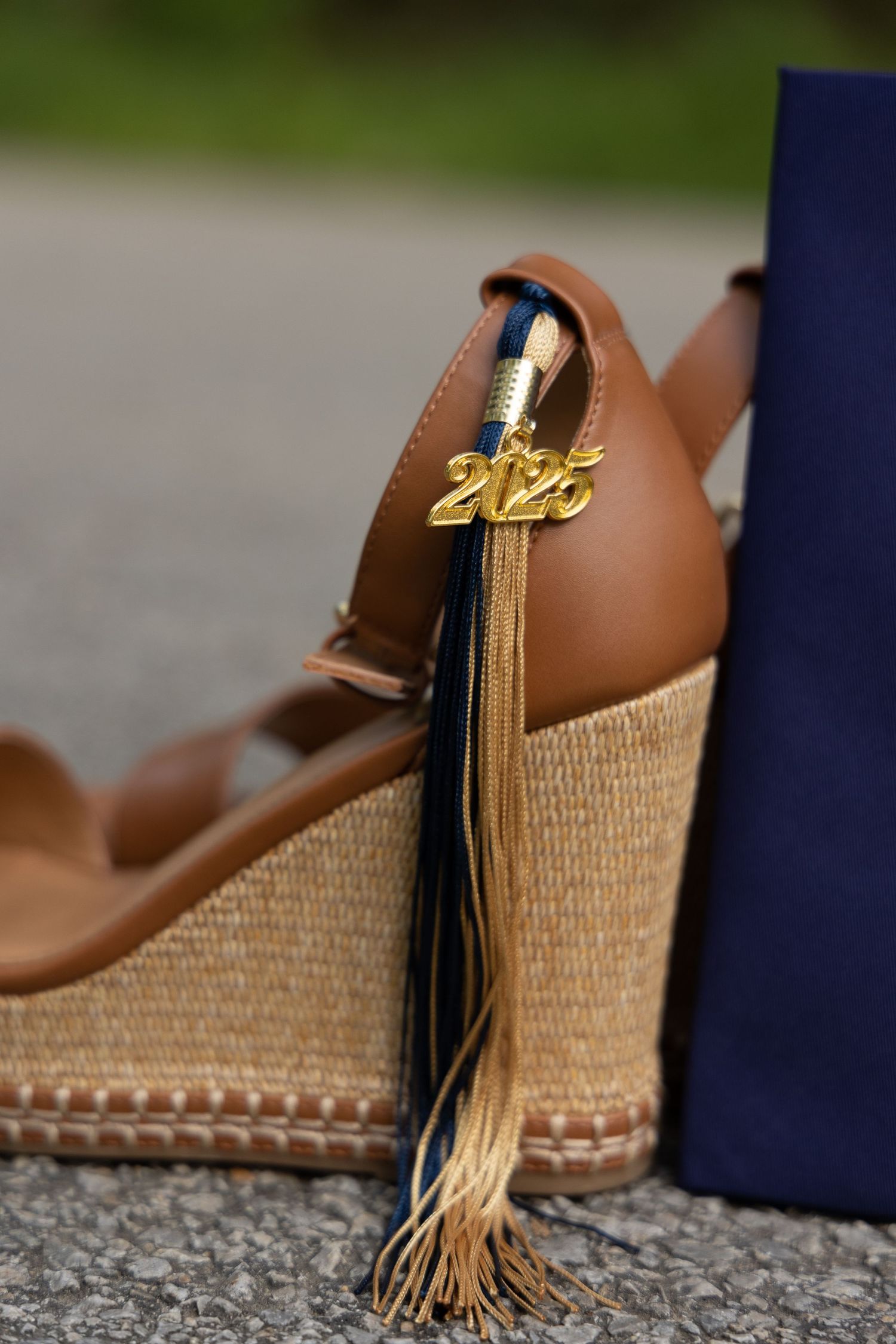A close-up of a graduation tassel with gold year charm resting on a brown leather and woven shoe.