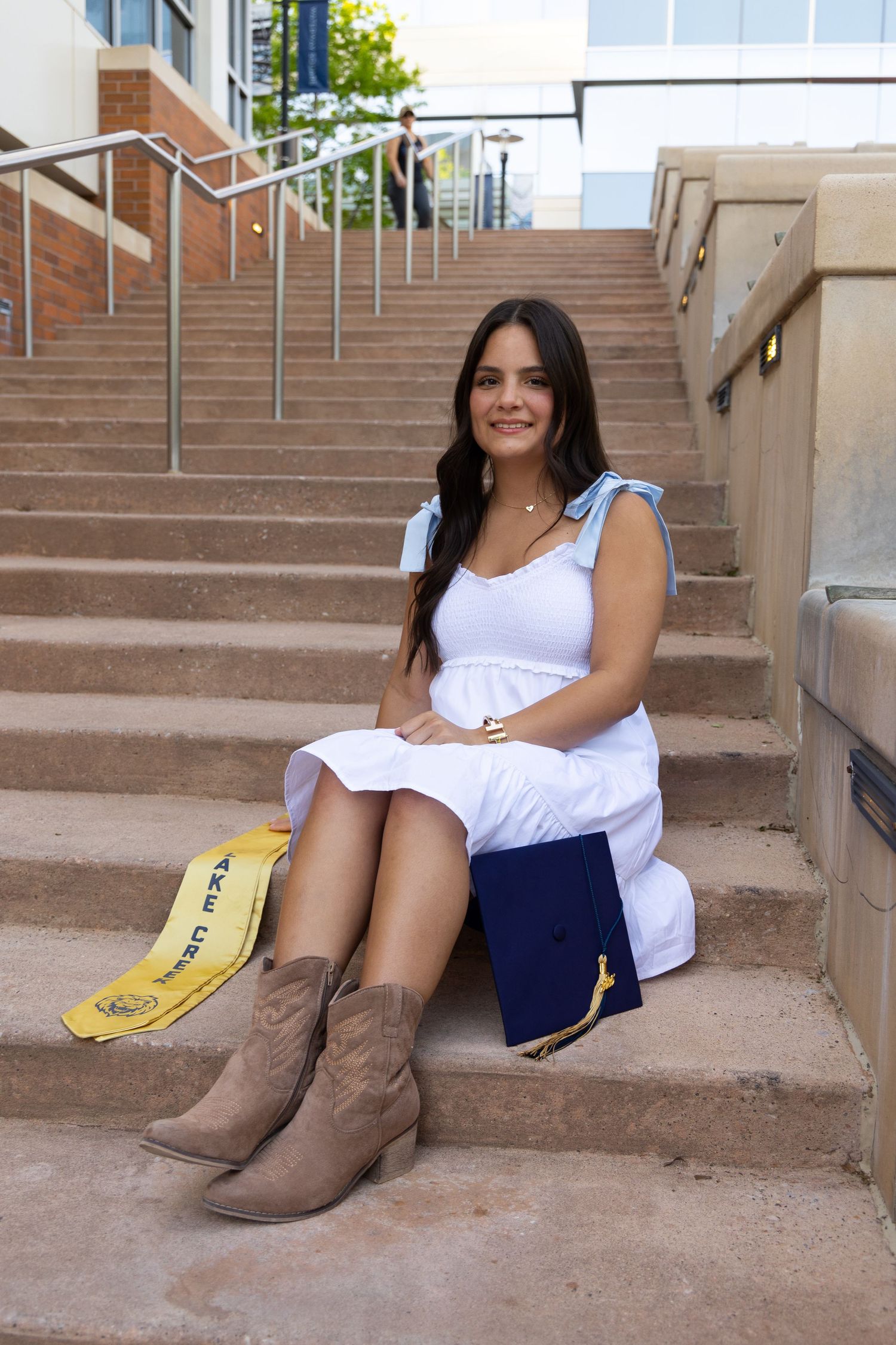 Young woman in white dress and boots sits on campus steps with graduation cap and sash, celebrating her achievement.