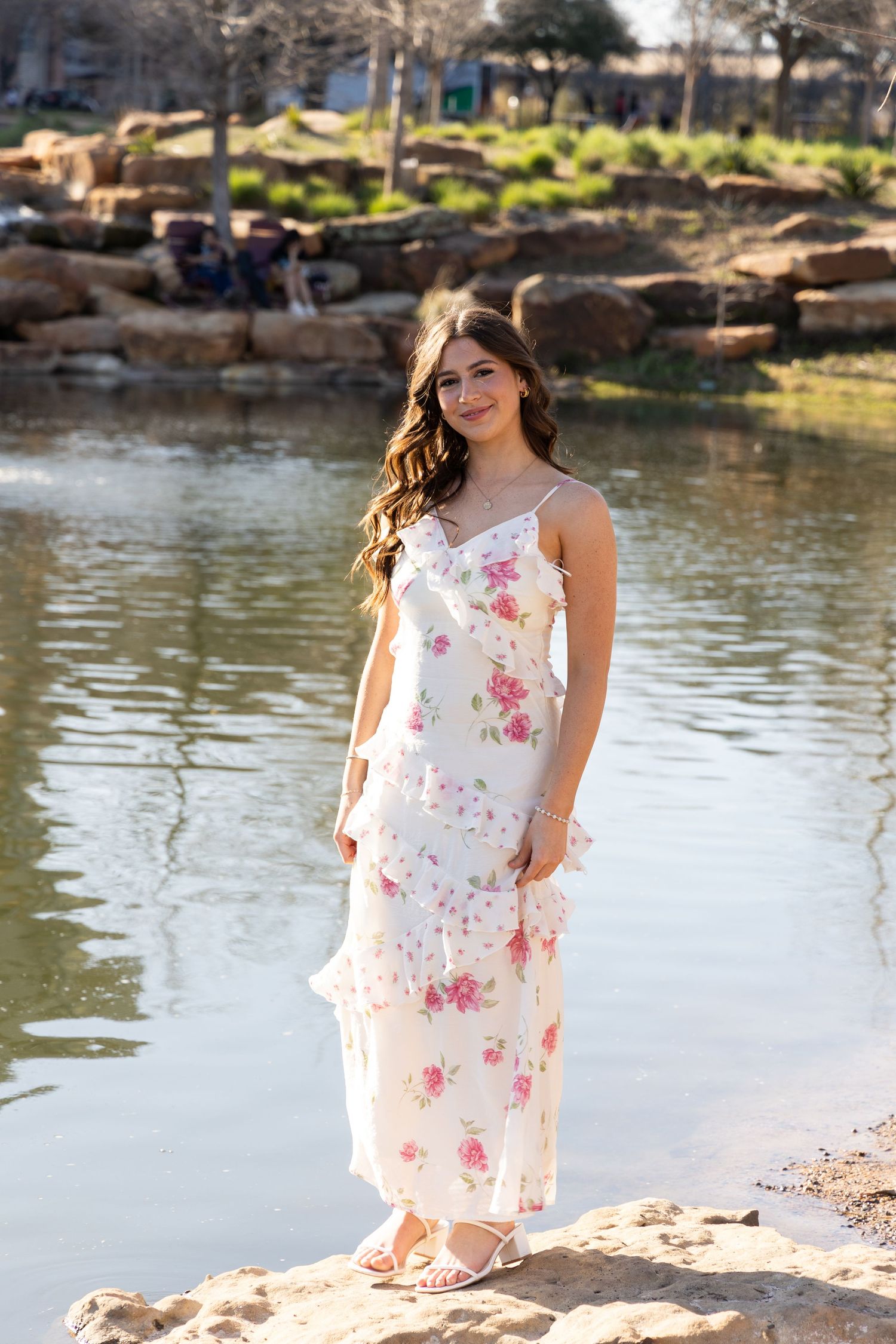 Young woman in white floral maxi dress posing by a serene pond on rocky shore, smiling outdoors.