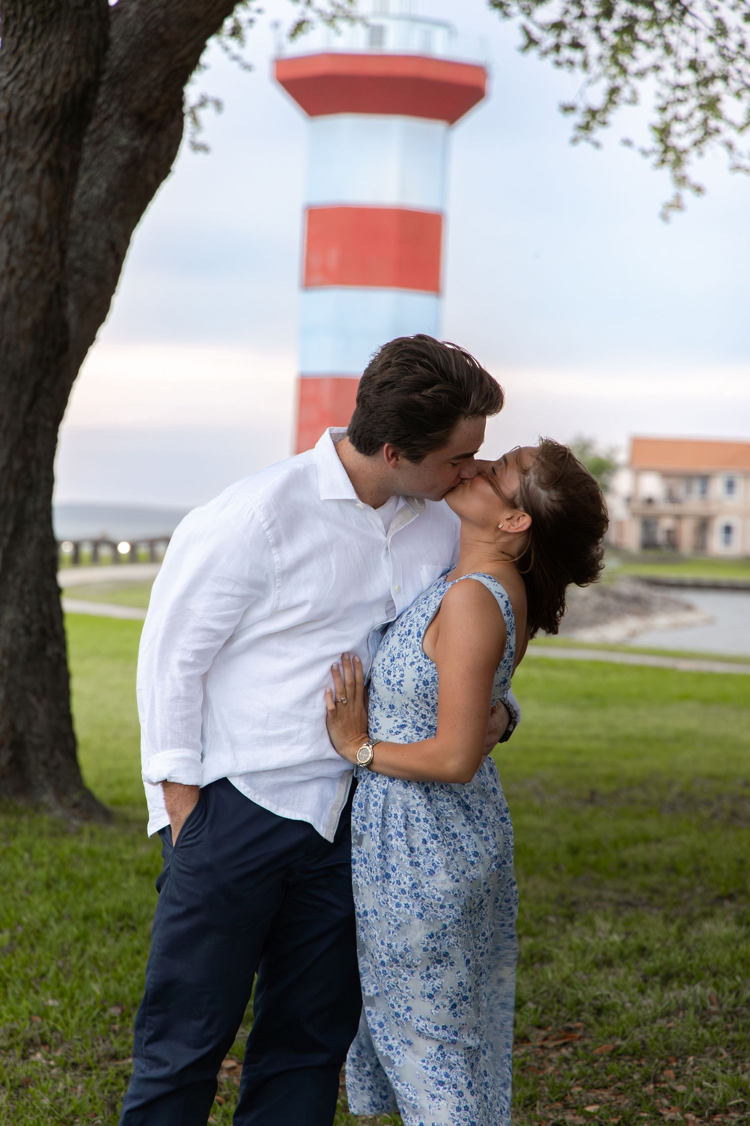 Couple sharing a romantic kiss near a red and white striped lighthouse on a green lawn.