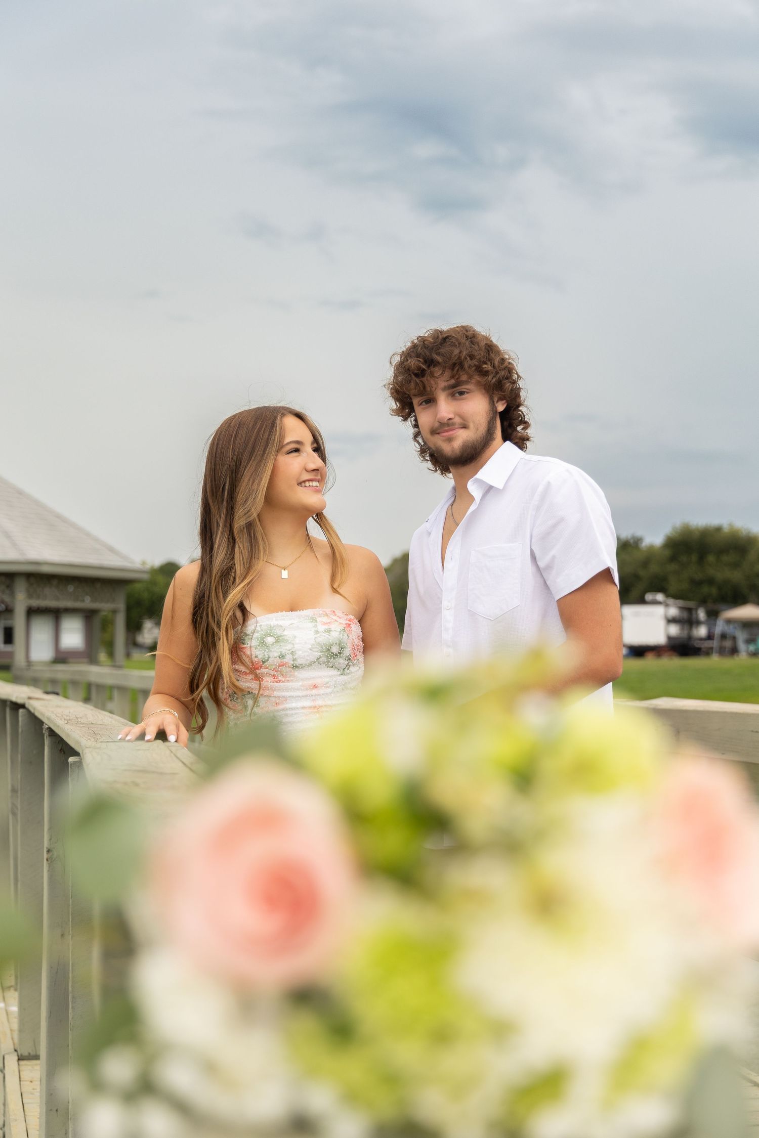 A young couple stands on a bridge surrounded by pink roses, smiling together under a cloudy sky.