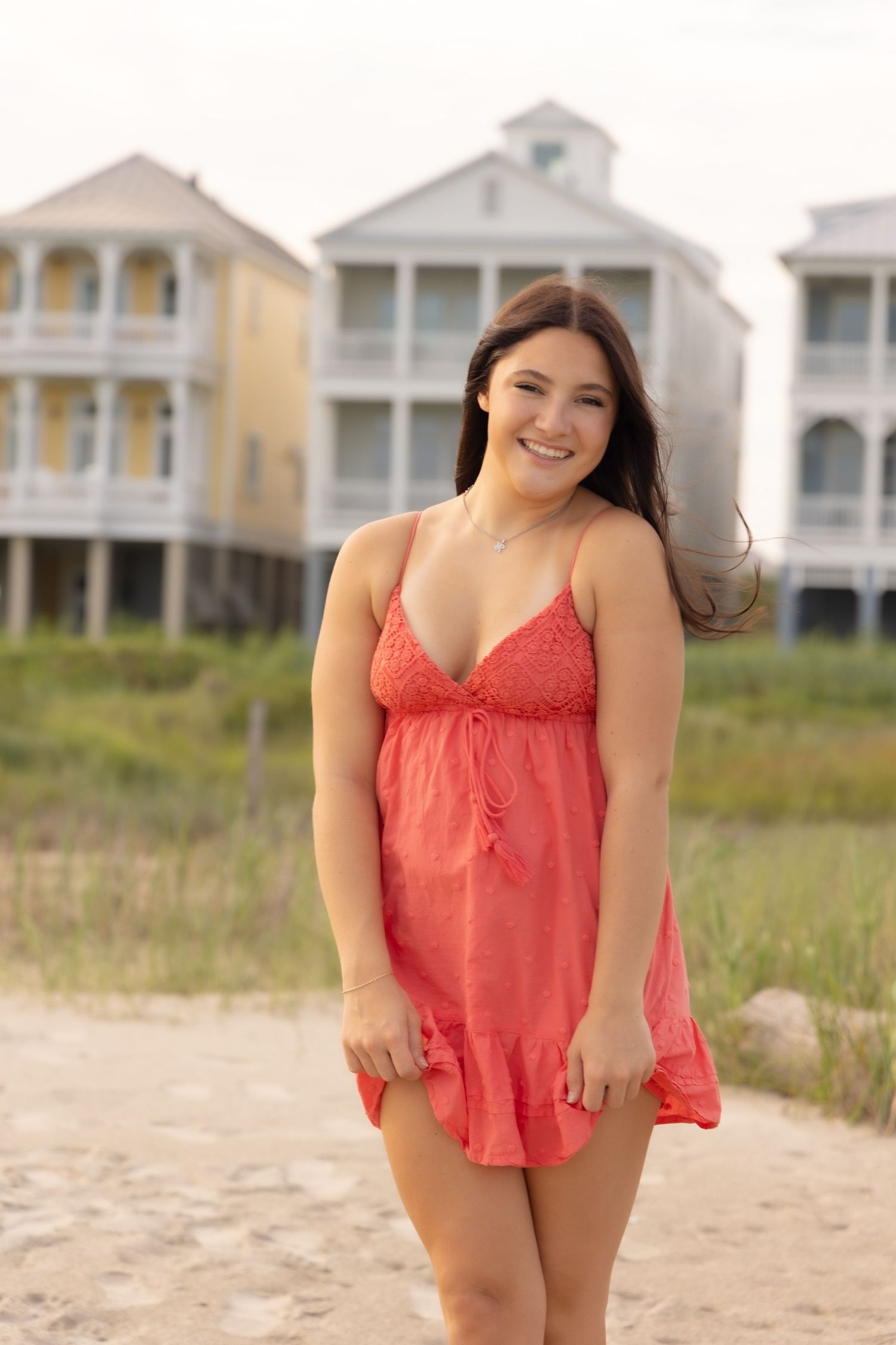 Happy young woman in red sundress smiling on sandy beach with colorful beach houses in background.