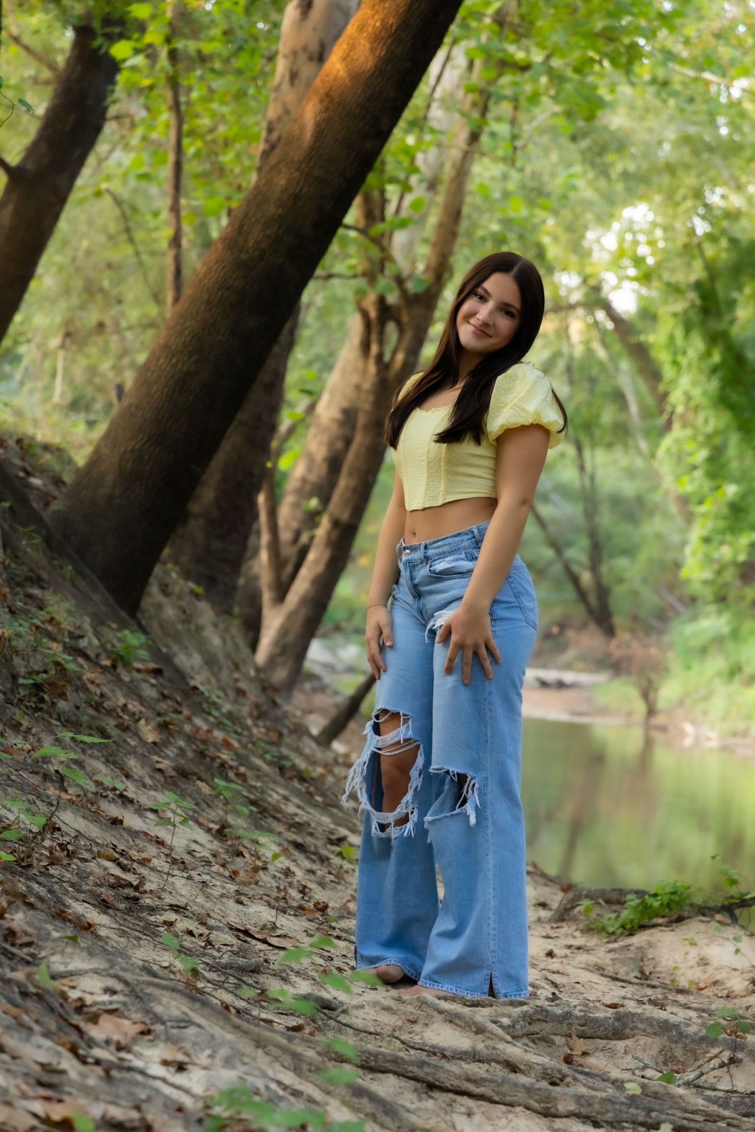 Young woman in yellow crop top and ripped blue jeans posing on a forest trail near a creek.