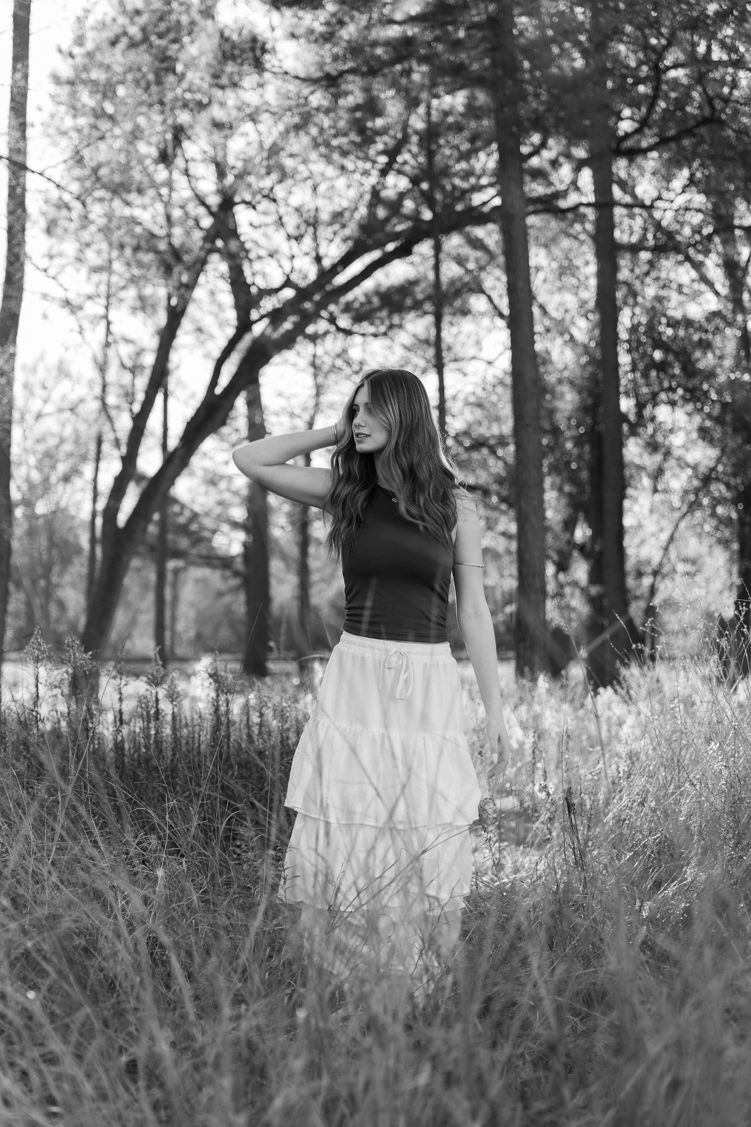 Black and white photo of a young woman in a white skirt standing in tall grass among trees.