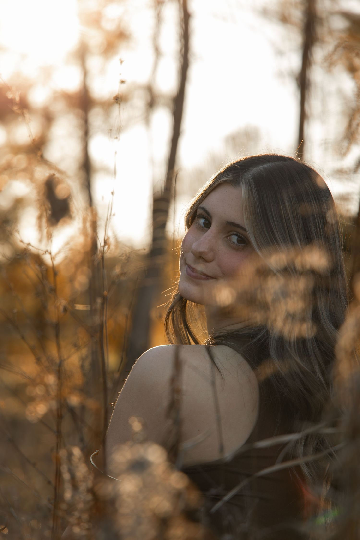 Young woman with long brown hair smiling softly among golden autumn foliage in warm sunset light.