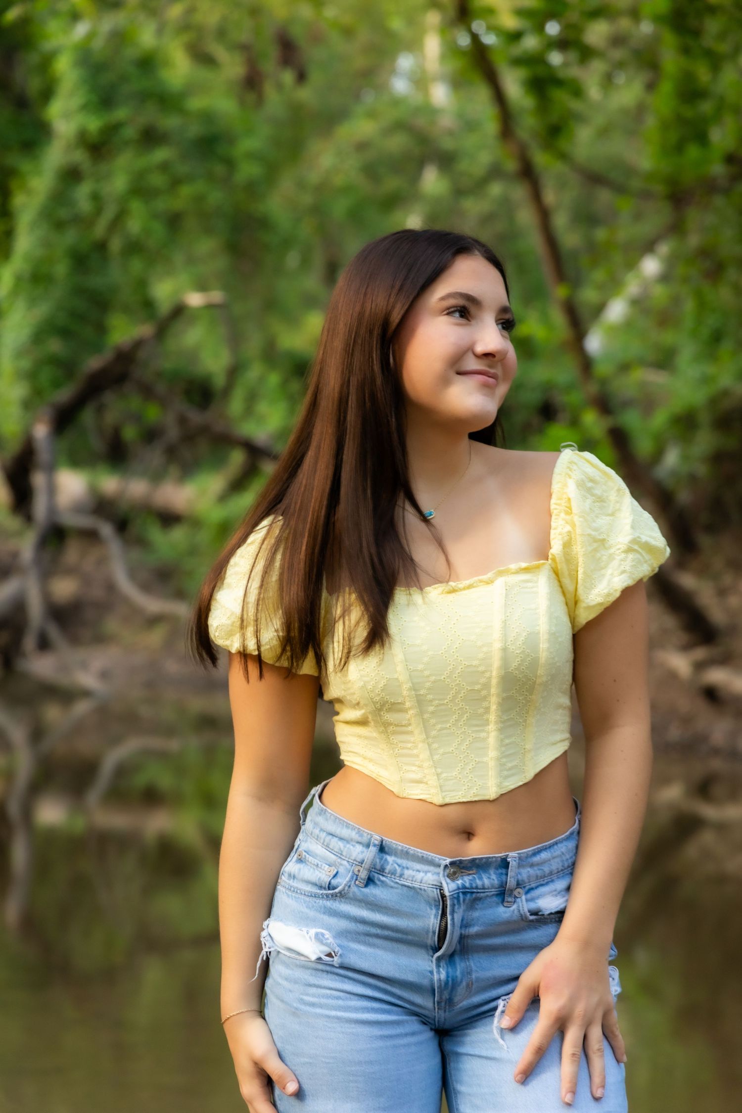 Young woman in yellow crop top and ripped jeans smiling outdoors with lush green trees in background.