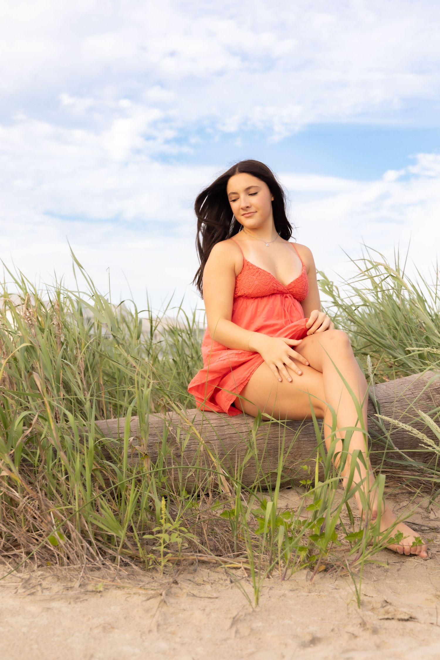 Young woman in red dress sitting on driftwood among beach grass with cloudy sky backdrop.