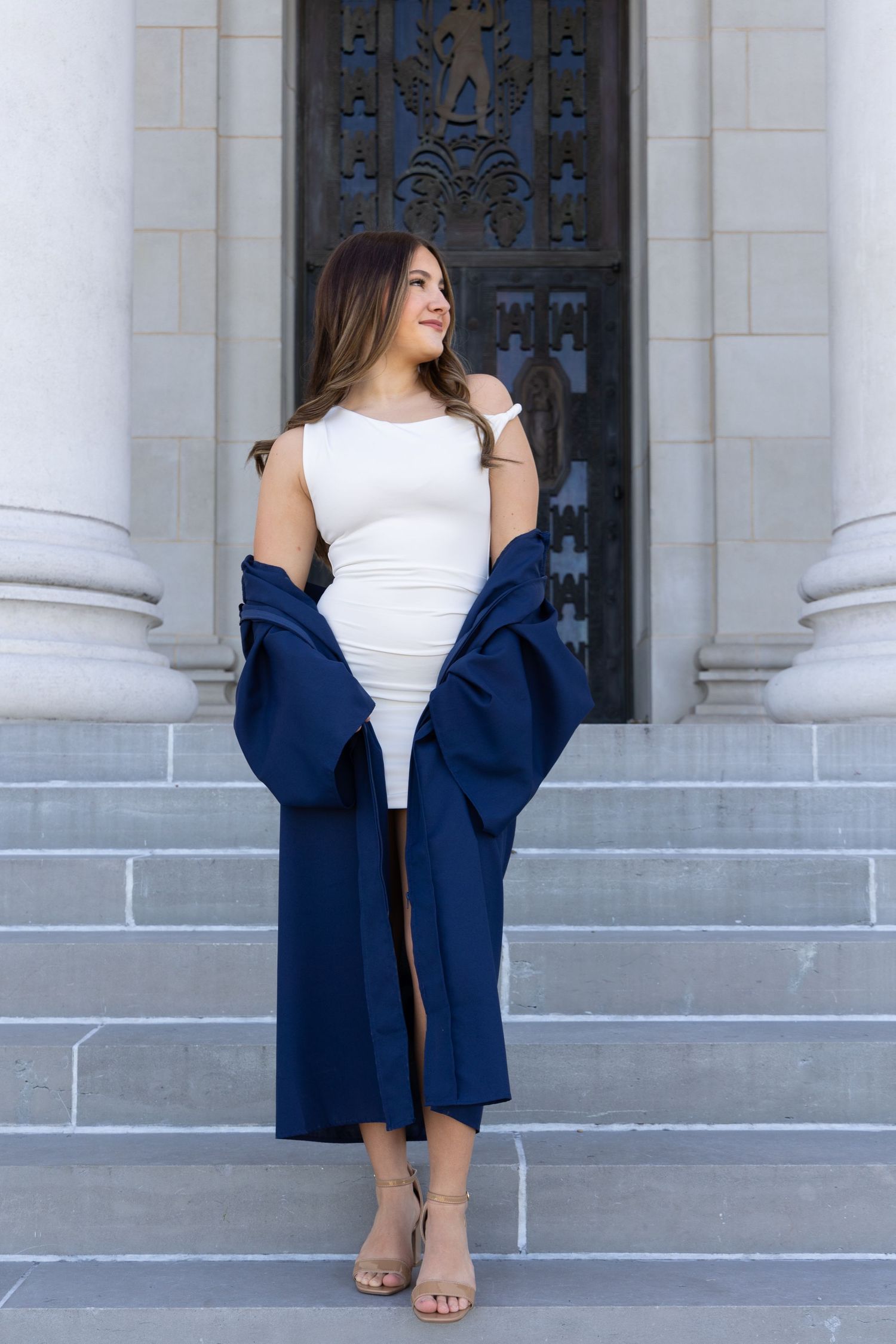 Young woman in white dress and navy blue coat poses gracefully on stone steps before ornate building doors.