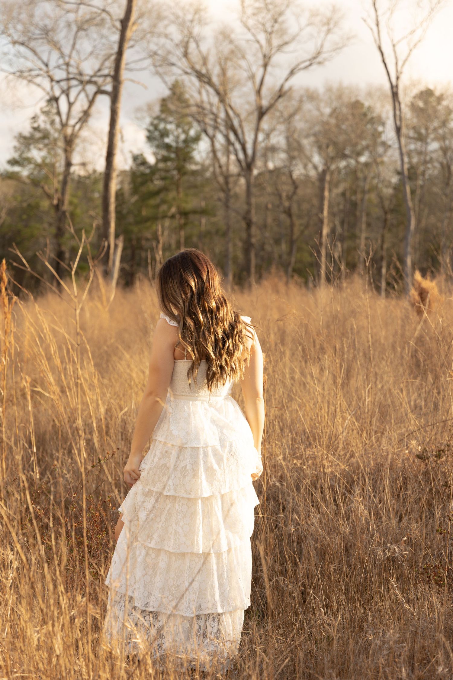 Woman in white tiered dress walking through tall grass at golden hour with trees in distance.