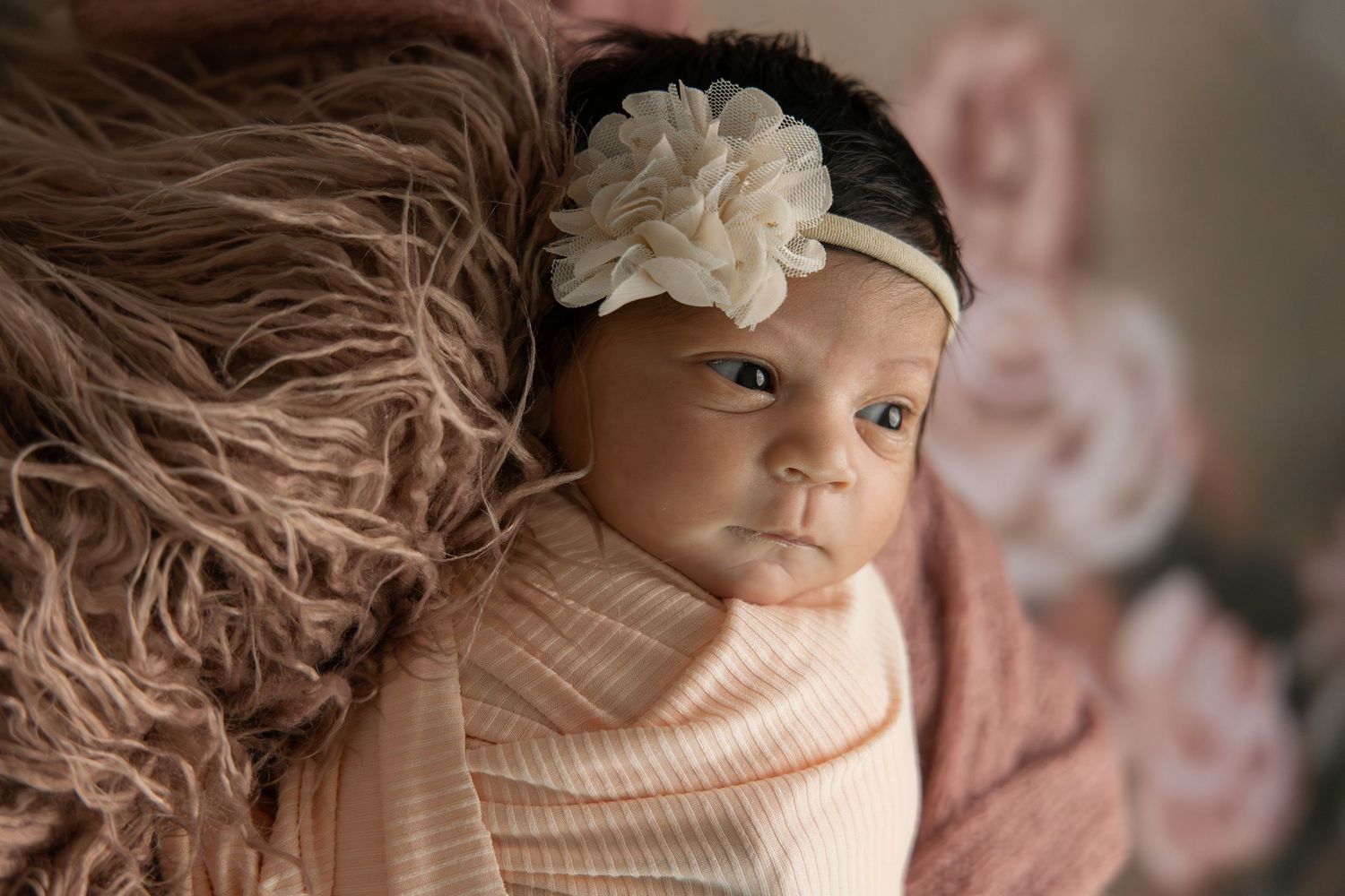 Newborn baby girl swaddled in peach wrap with cream floral headband, resting on pink fluffy blanket.