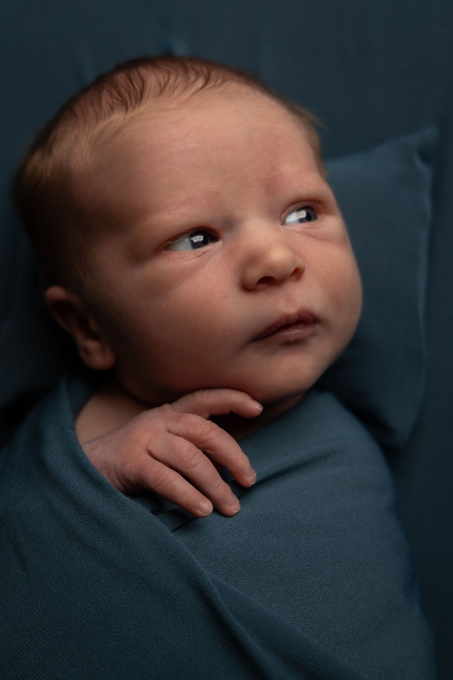 Newborn baby wrapped in teal swaddle blanket, alert with open eyes, photographed in soft studio lighting.
