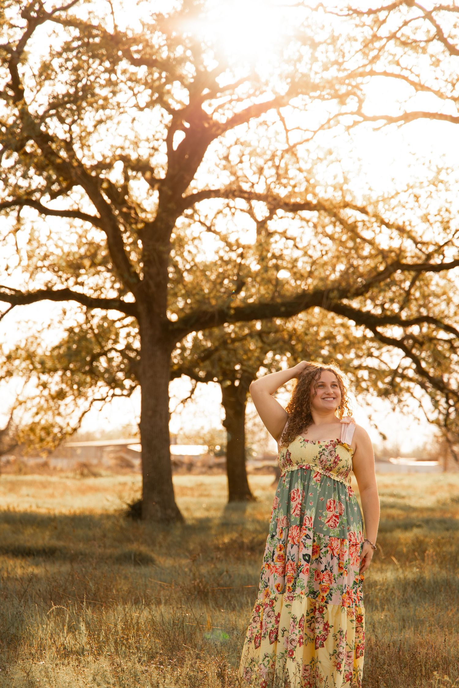 Woman in floral dress standing in sunlit field with large oak trees glowing in warm golden hour light.