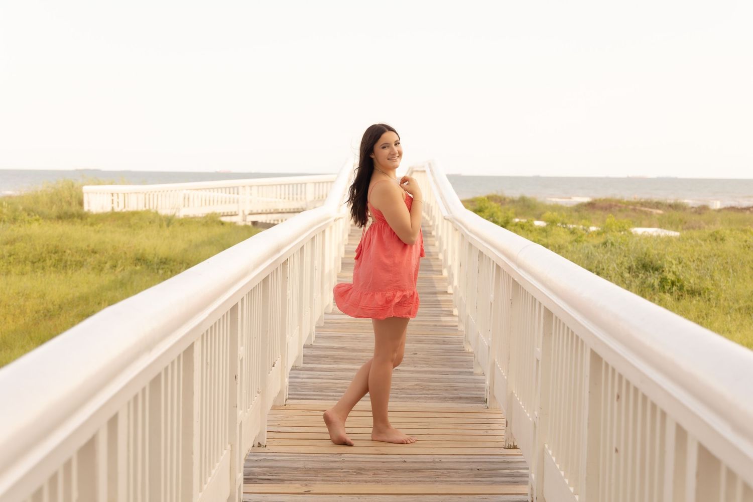 Young woman in coral dress standing on a white wooden boardwalk leading to the beach on a sunny day.