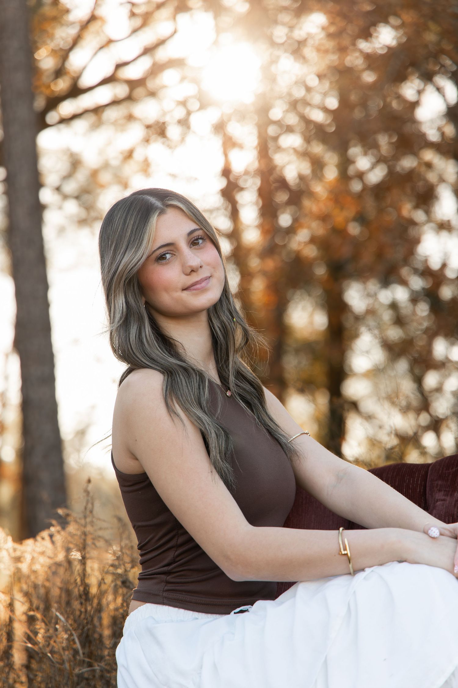 Young woman with long wavy hair in brown top and white skirt sitting outdoors among autumn trees in golden light.