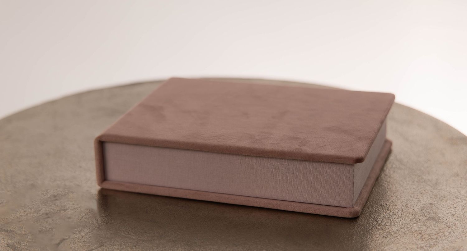 A dusty pink suede hardcover book box resting on a round stone surface against a neutral background.