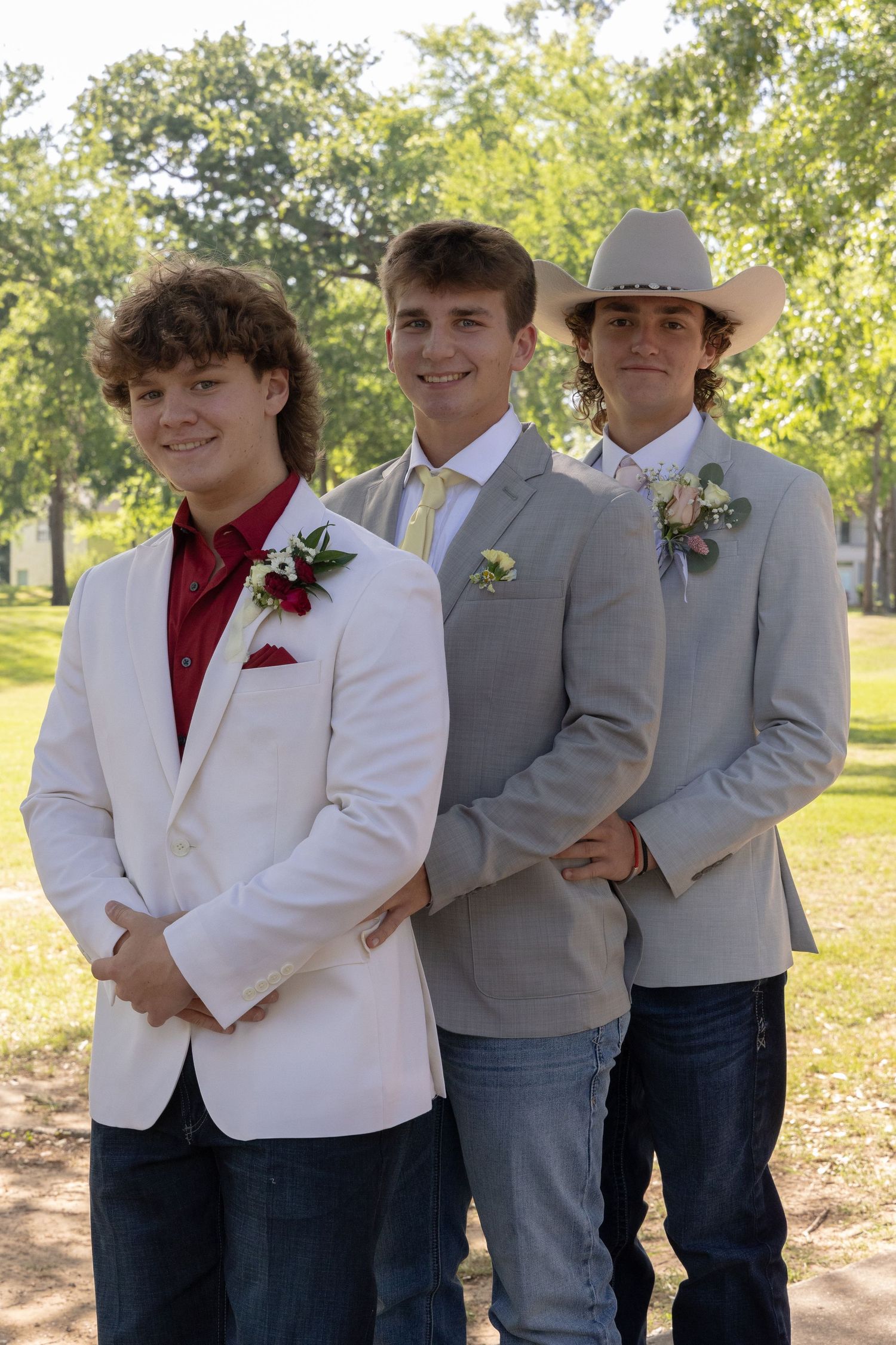 Three teenage boys in blazers and boutonnieres pose outdoors for prom photos.