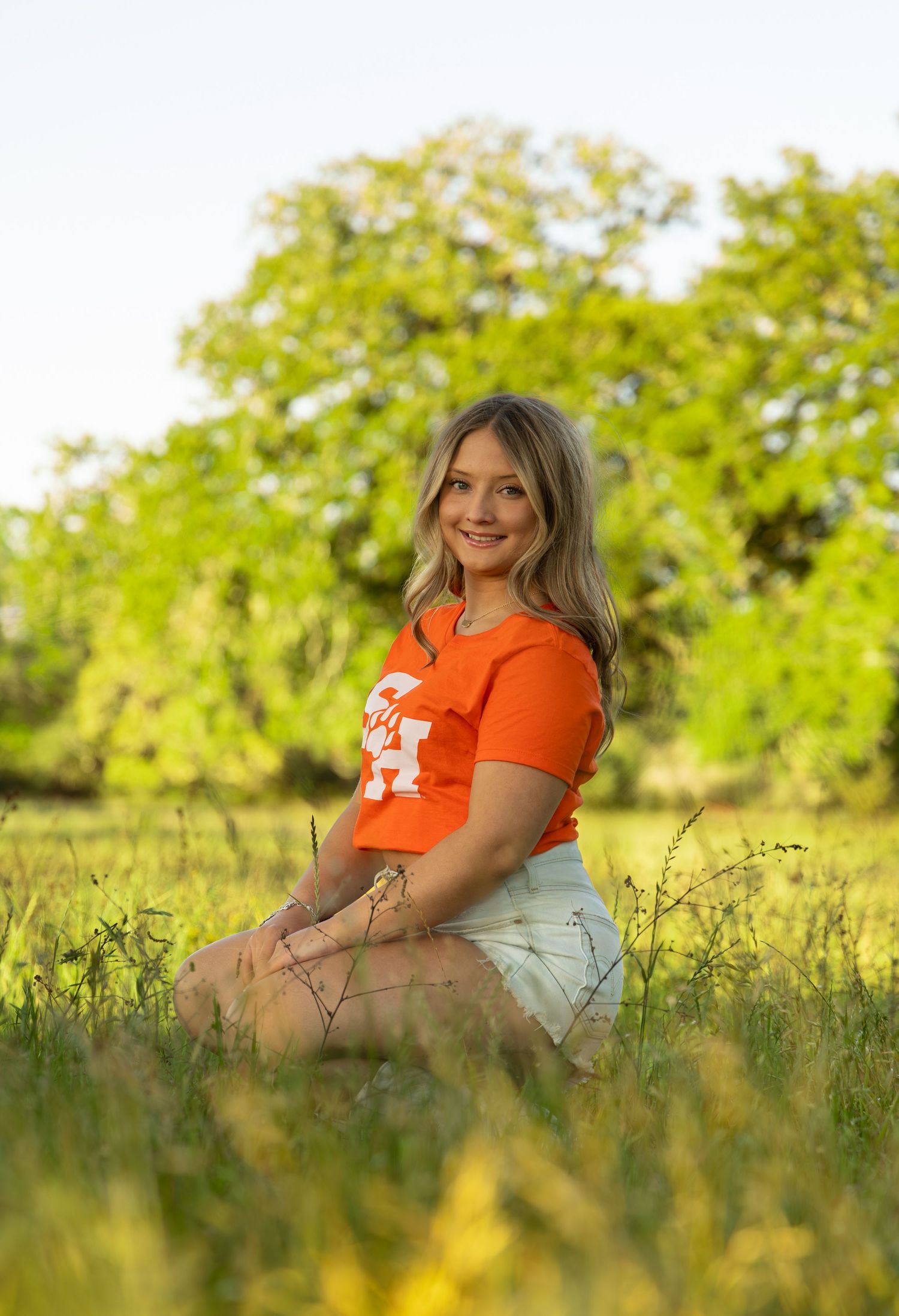 Young woman in orange university t-shirt sitting in green meadow, smiling outdoors during golden hour.