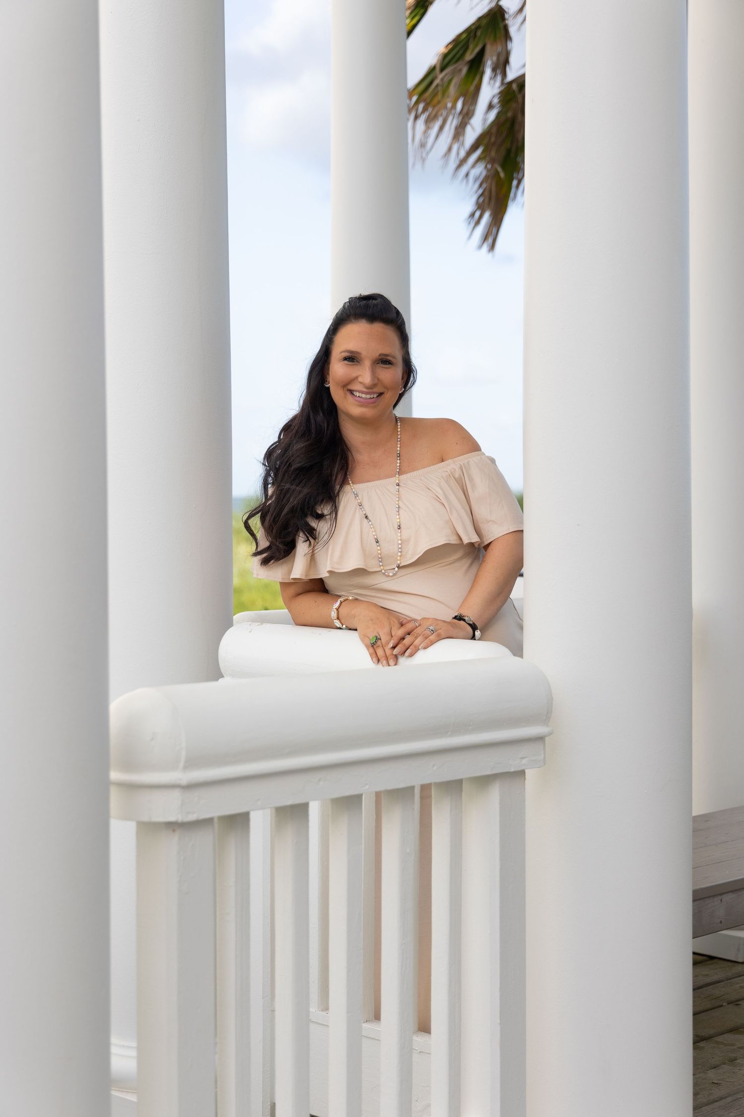 Smiling brunette woman in off-shoulder beige top leaning on white porch railing between columns with palm trees.