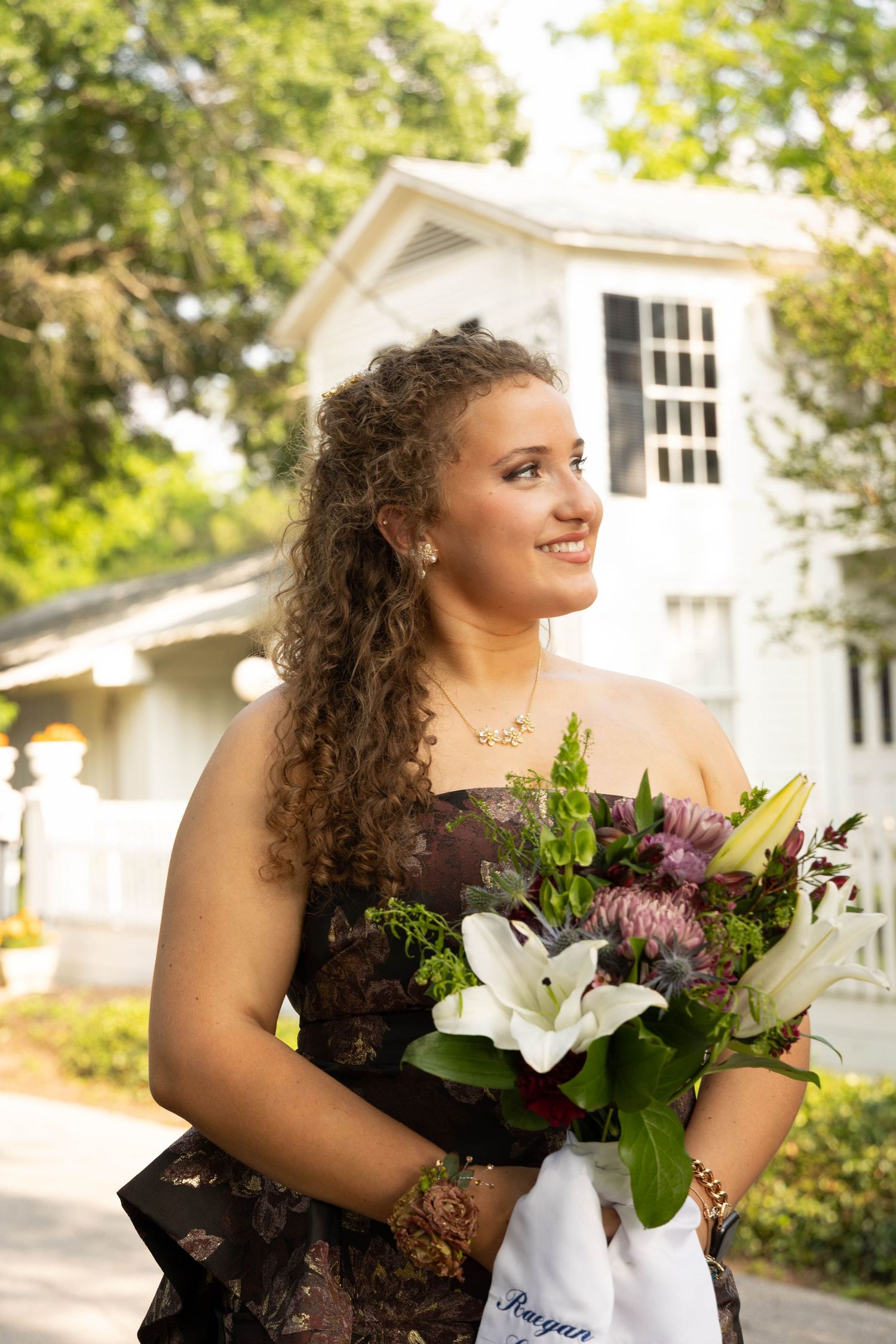 Young woman in floral gown holding bouquet of white lilies and purple flowers smiling outdoors near white house.
