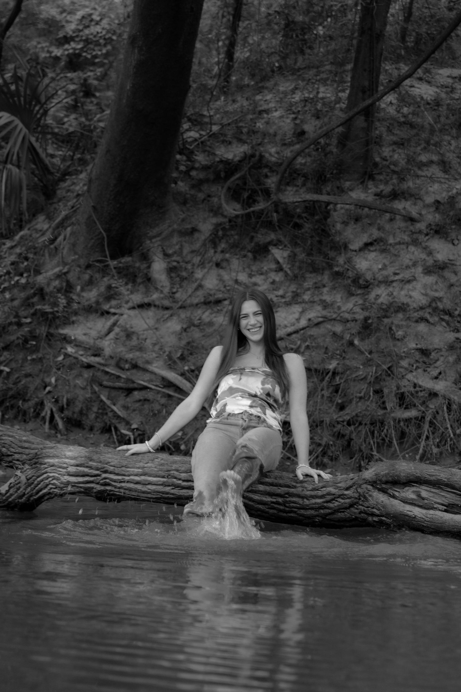 Young woman sitting on a fallen log in a forest creek, splashing water, black and white nature photography.