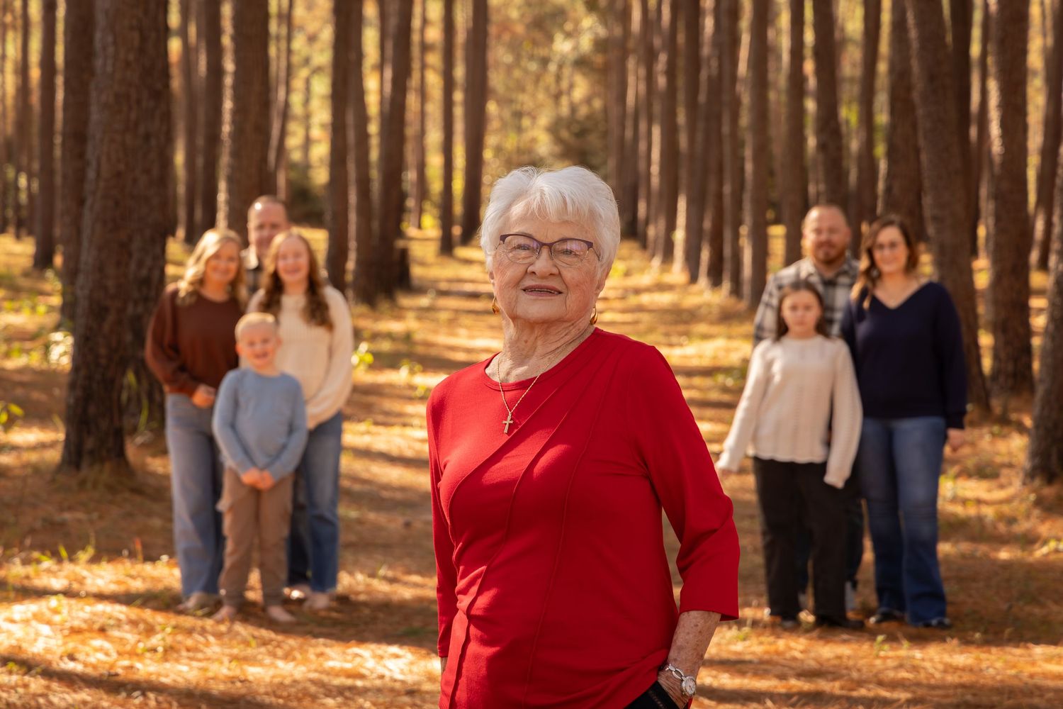 Elderly woman in red sweater smiles in pine forest with family group posed behind her in warm autumn light.