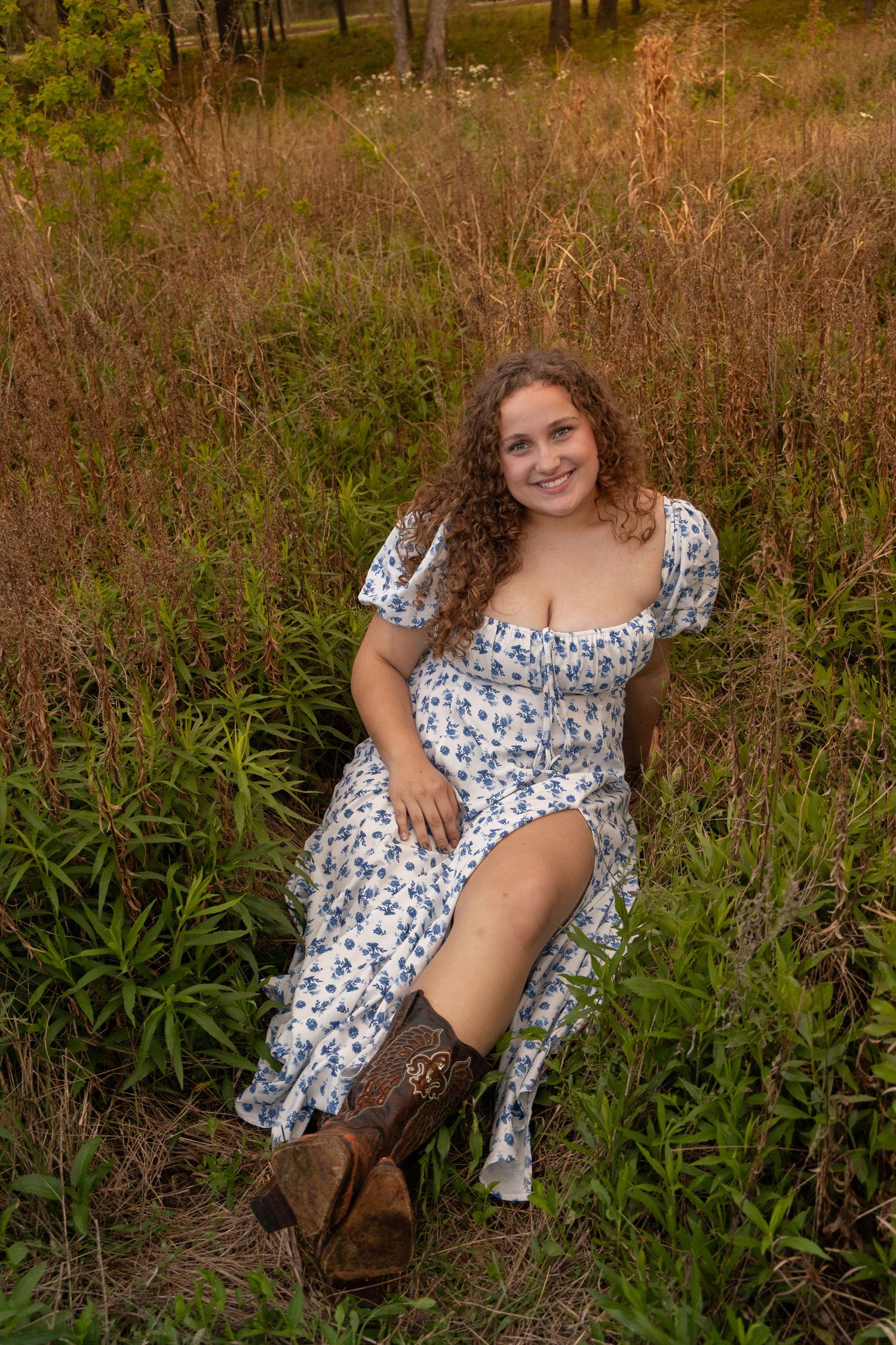 Young woman in floral dress and cowboy boots sitting in a lush green meadow, smiling at the camera.