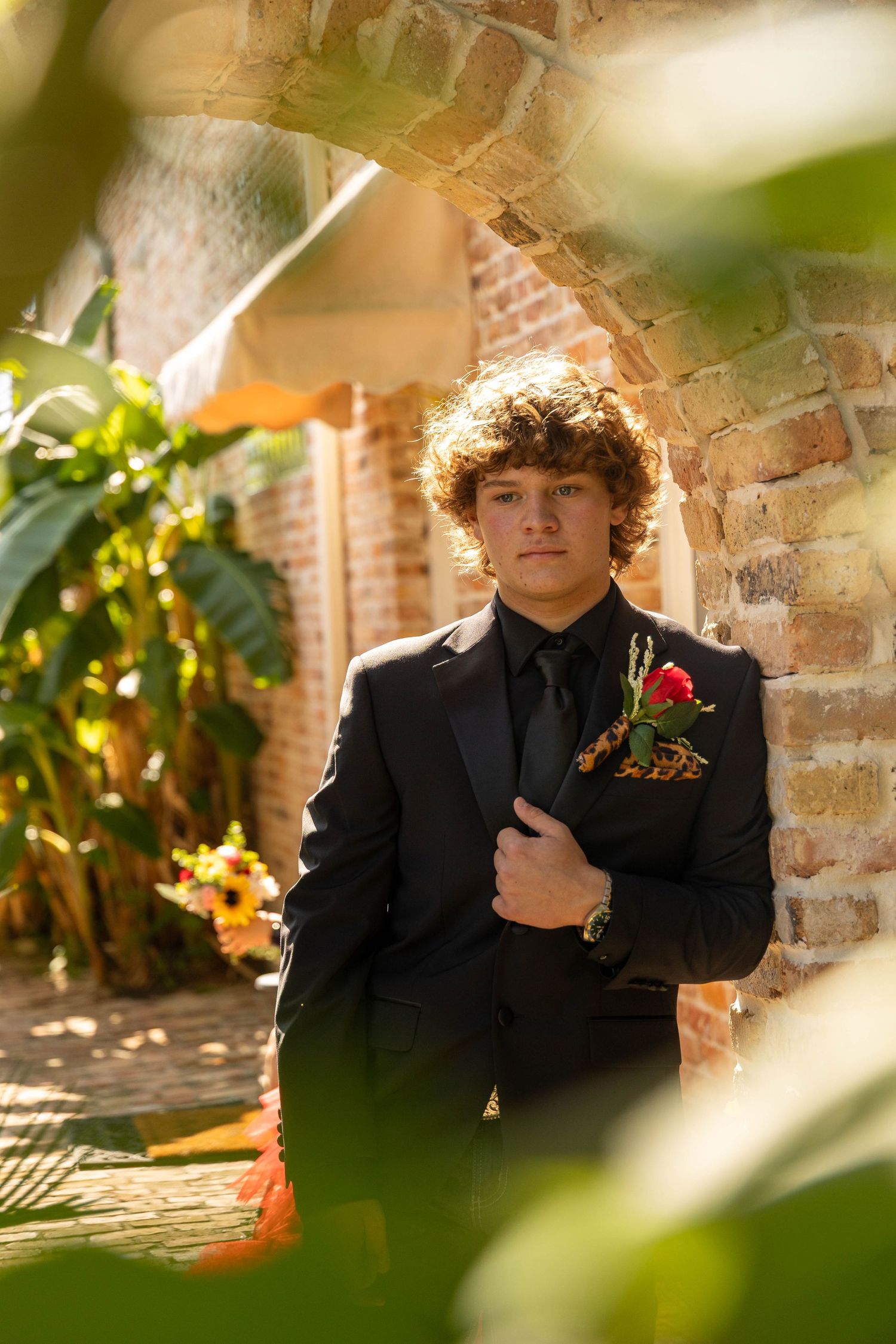 Young man in black suit with red rose boutonniere poses confidently against rustic brick wall surrounded by lush greenery.