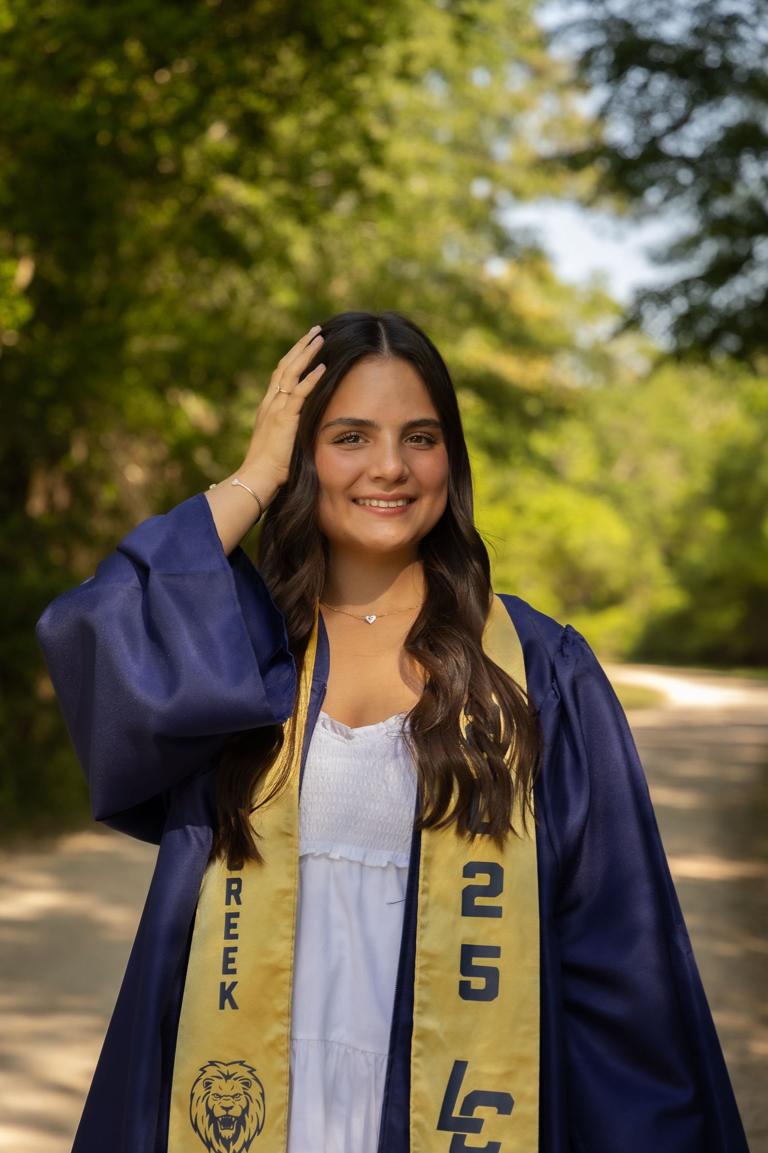 Smiling graduate in navy blue cap and gown with gold stole poses outdoors among green trees on graduation day.