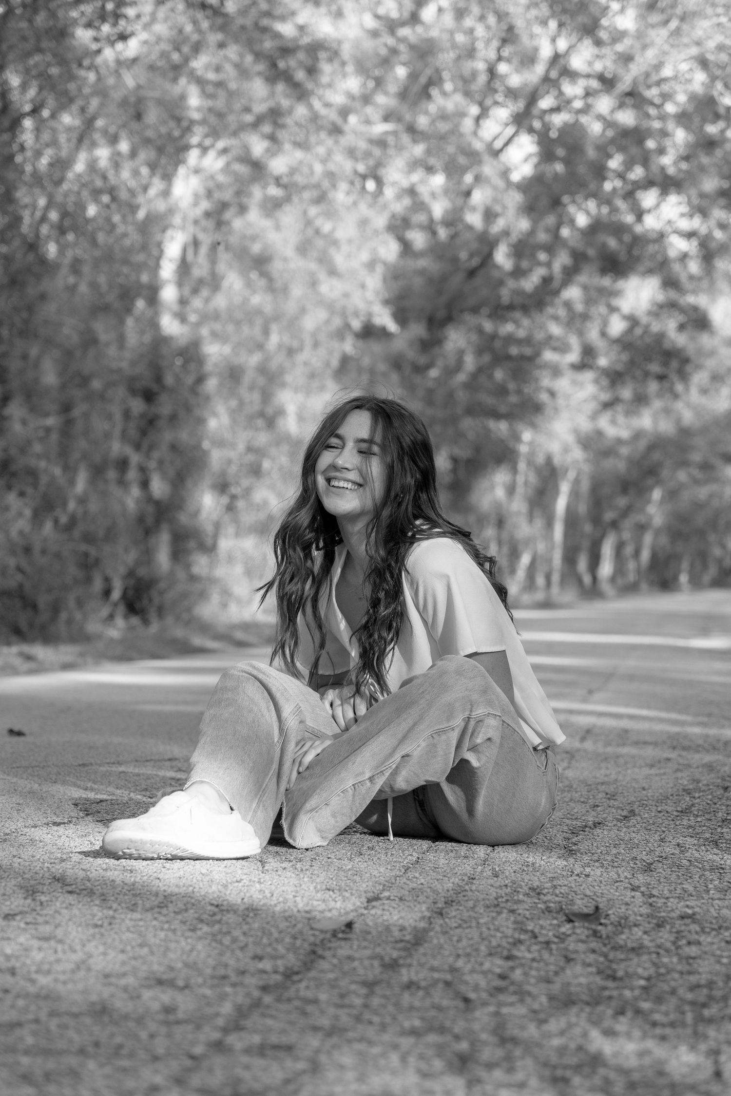 Black and white photo of person with long wavy hair sitting on ground outdoors wearing white top and casual pants.