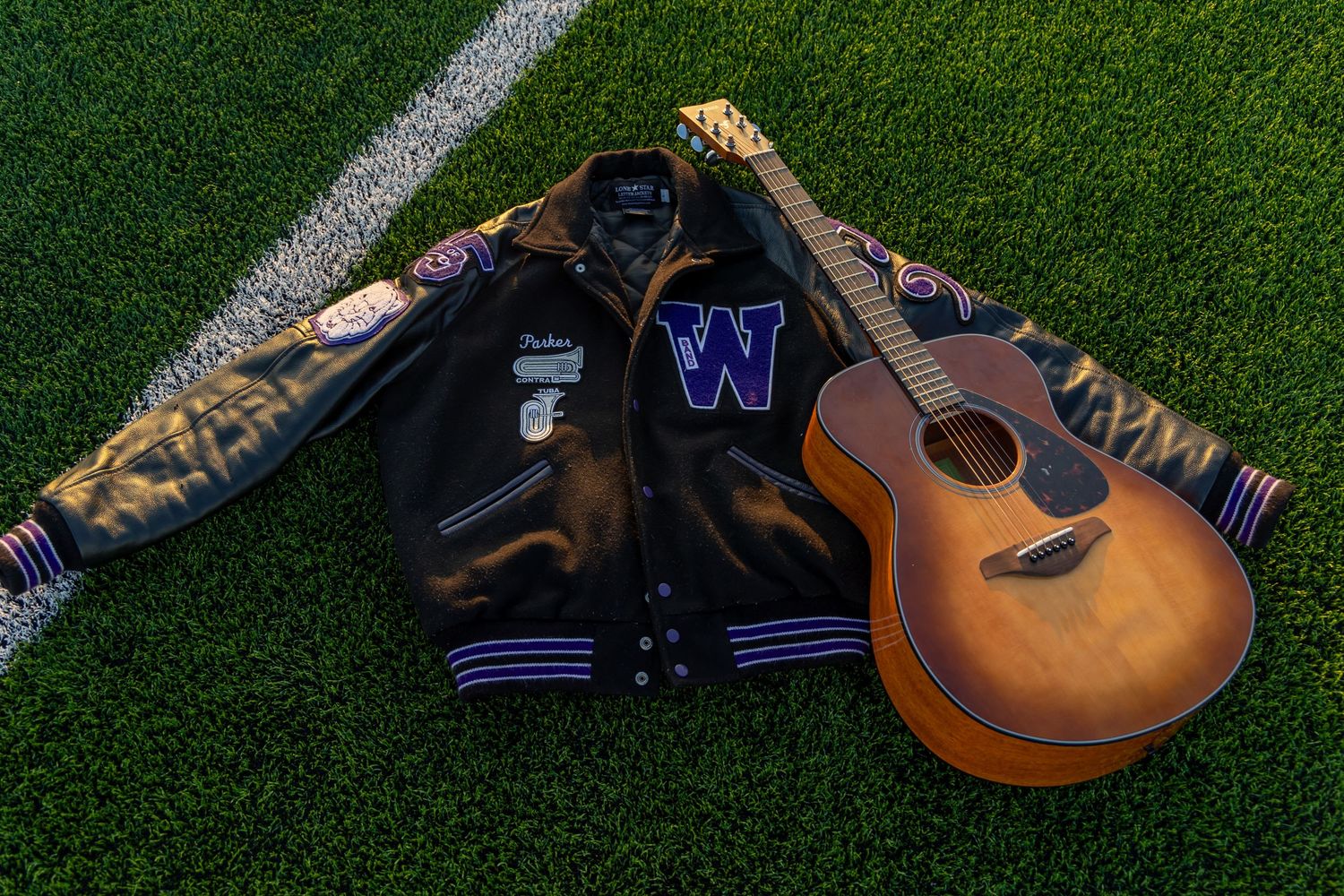 A varsity letterman jacket with 'W' logo and an acoustic guitar laid on green turf with white field markings.