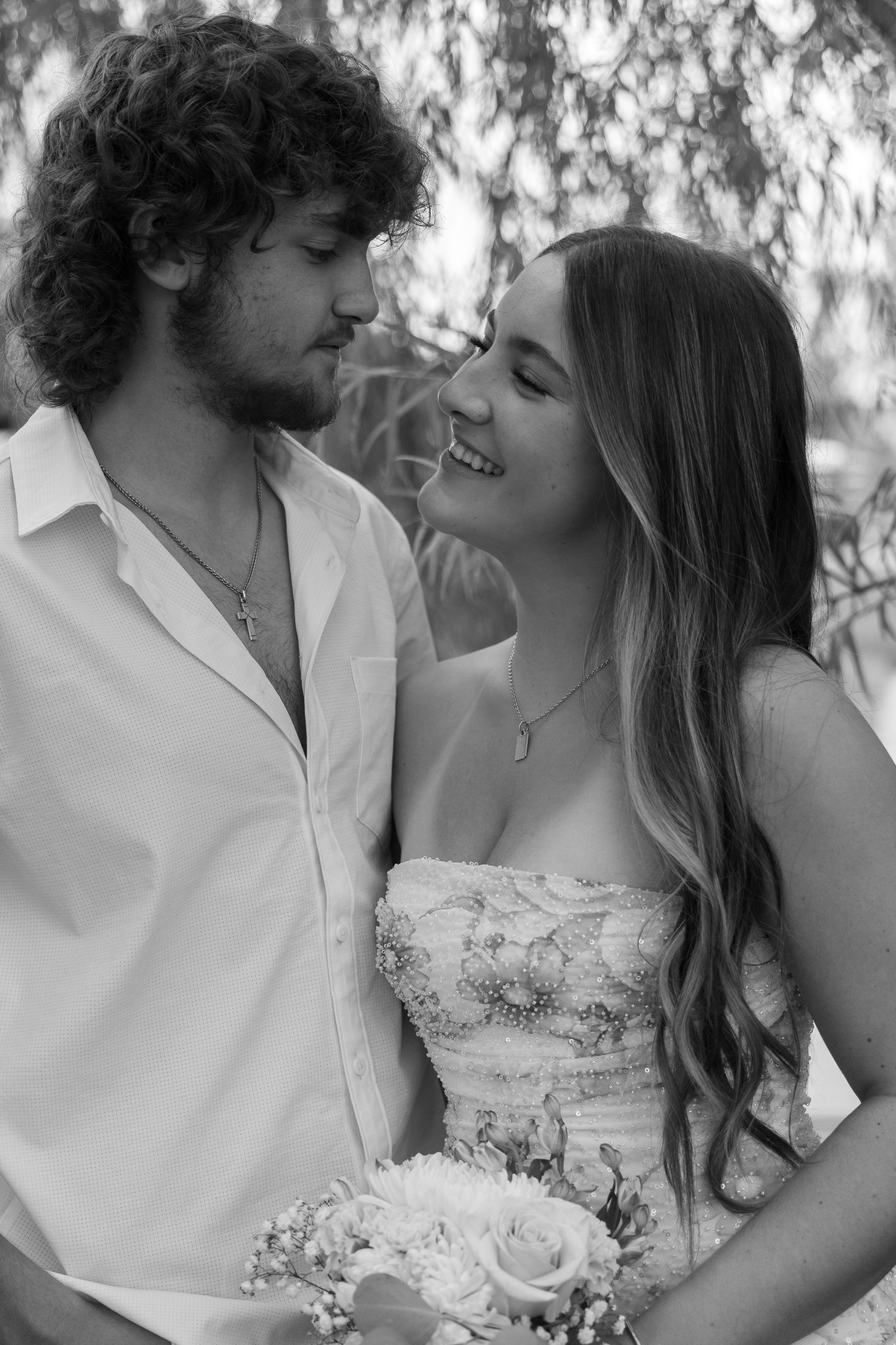 Black and white photo of a smiling bride holding flowers, gazing at a curly-haired groom in a white shirt outdoors.
