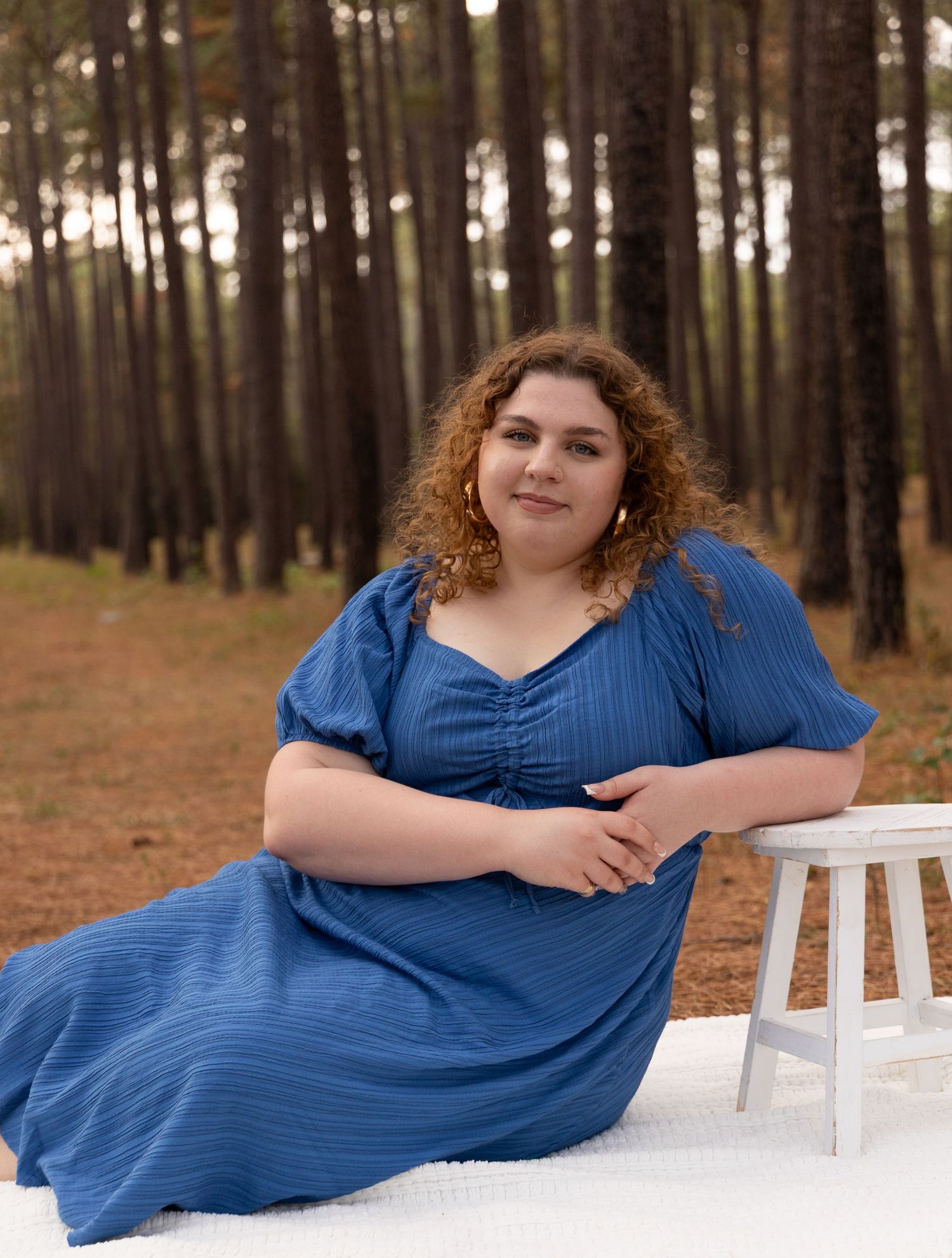 Woman in blue maxi dress poses gracefully in pine forest, sitting on white blanket beside wooden stool.