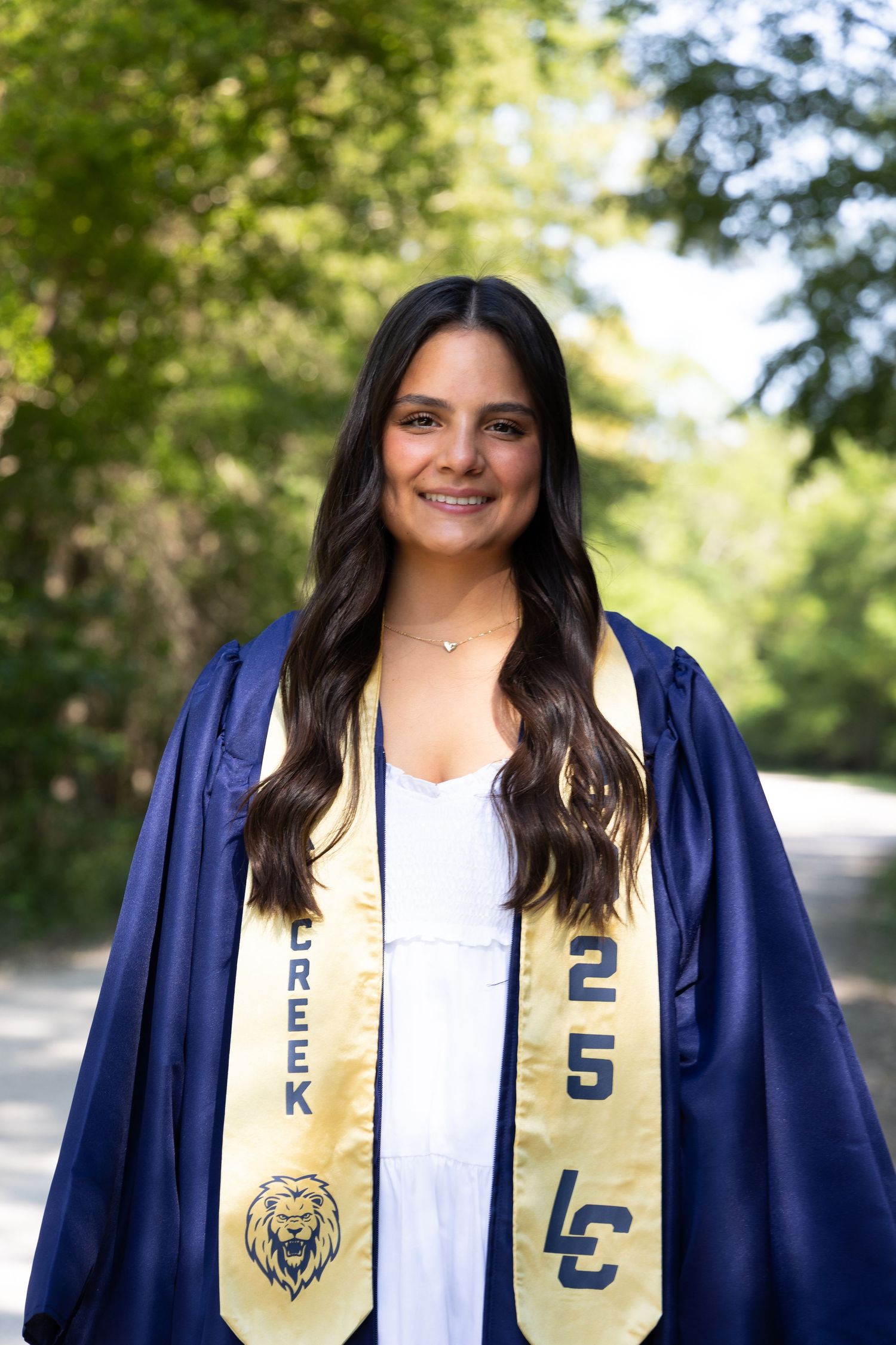 Smiling young woman in navy blue graduation gown and gold LC stole with lion mascot, posing outdoors among green trees.