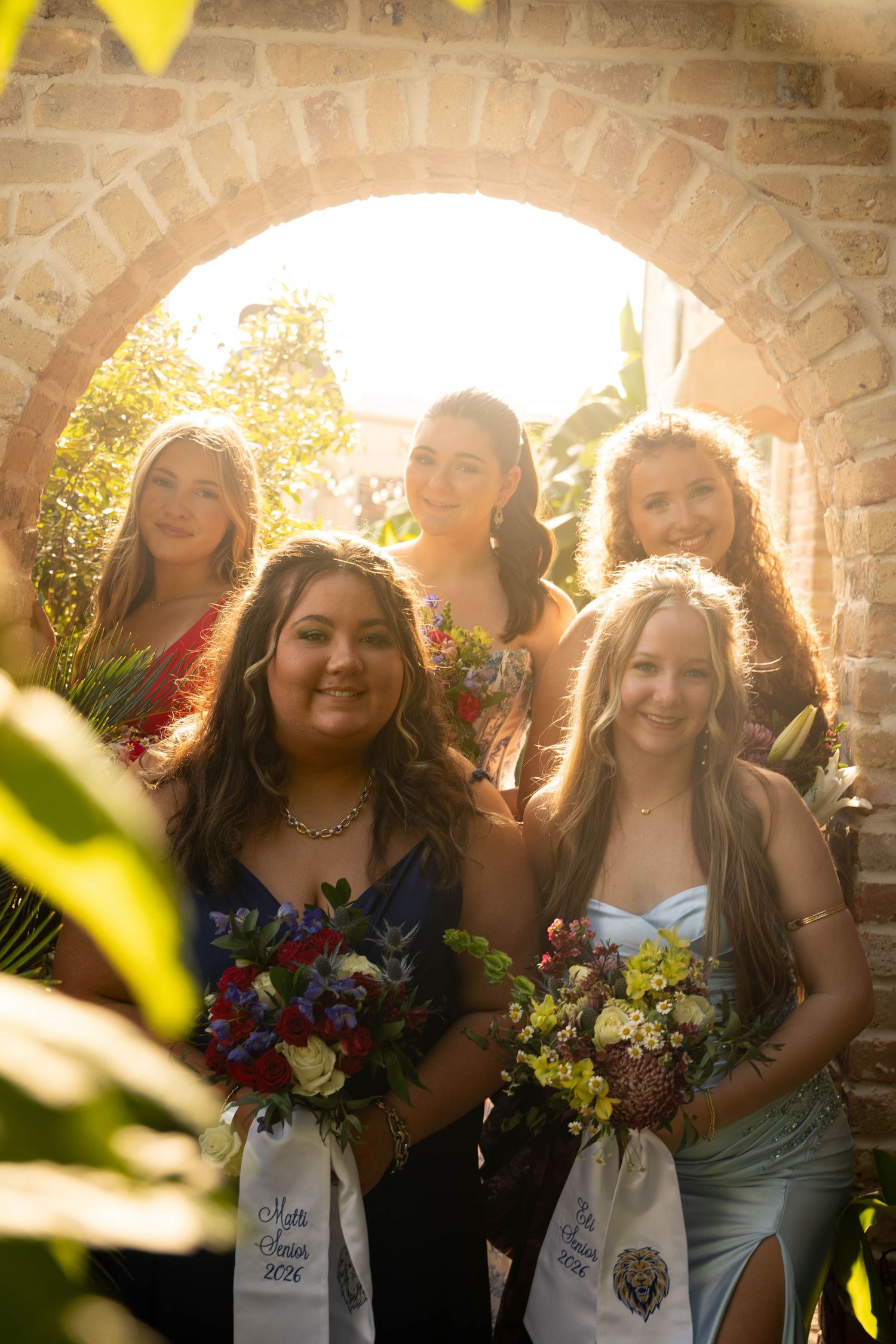 Group of five young women in formal dresses holding bouquets posing inside sunlit circular brick archway.