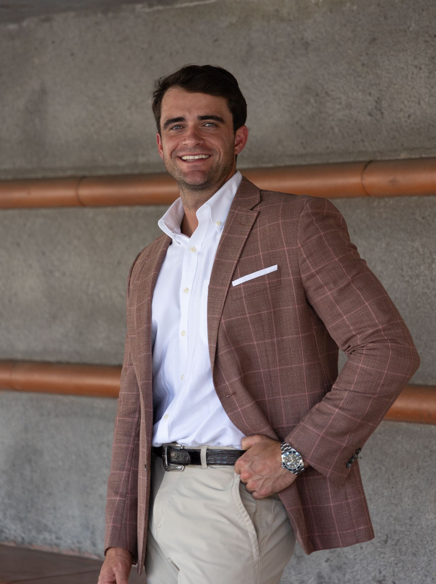 Smiling man in brown plaid blazer and white shirt poses confidently indoors near copper pipes.