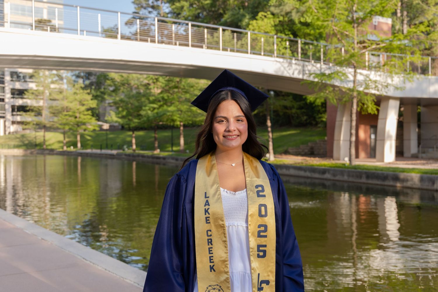 Smiling graduate in navy cap and gown with 2025 stole poses outdoors near a scenic pond and bridge.