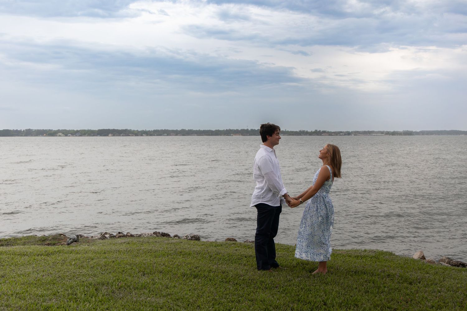 Romantic couple holding hands by the waterfront on a cloudy day near a calm bay.
