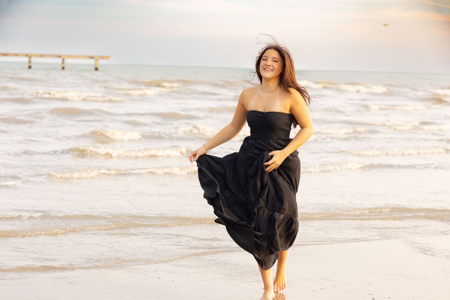 Woman in black strapless dress walking on beach with ocean waves in background at sunset.