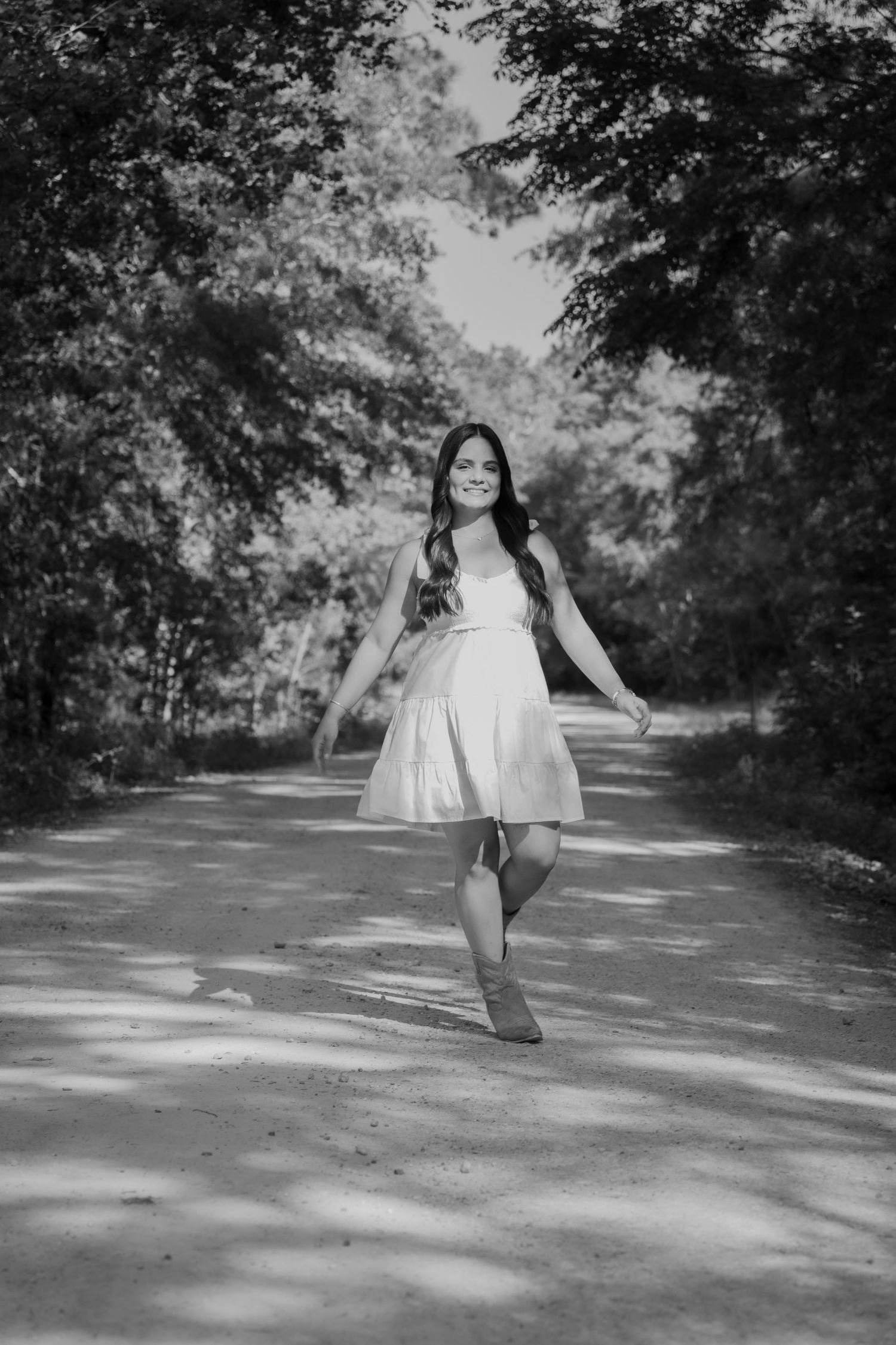 A figure in a white summer dress spins gracefully on a sunlit tree-lined path in a black and white photograph.