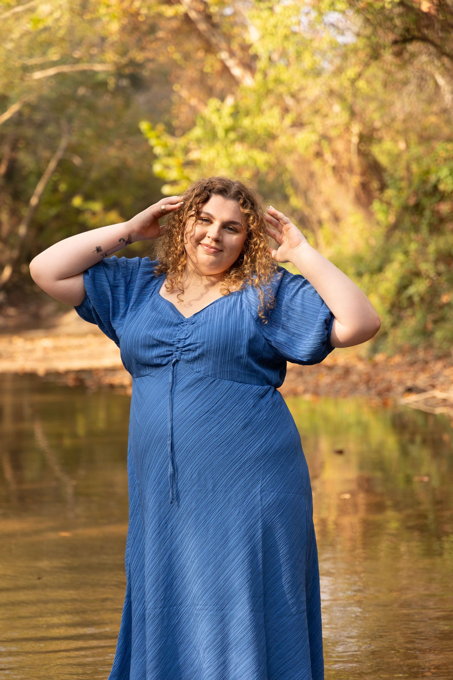 Plus-size woman in blue maxi dress posing by a serene creek surrounded by lush green trees.