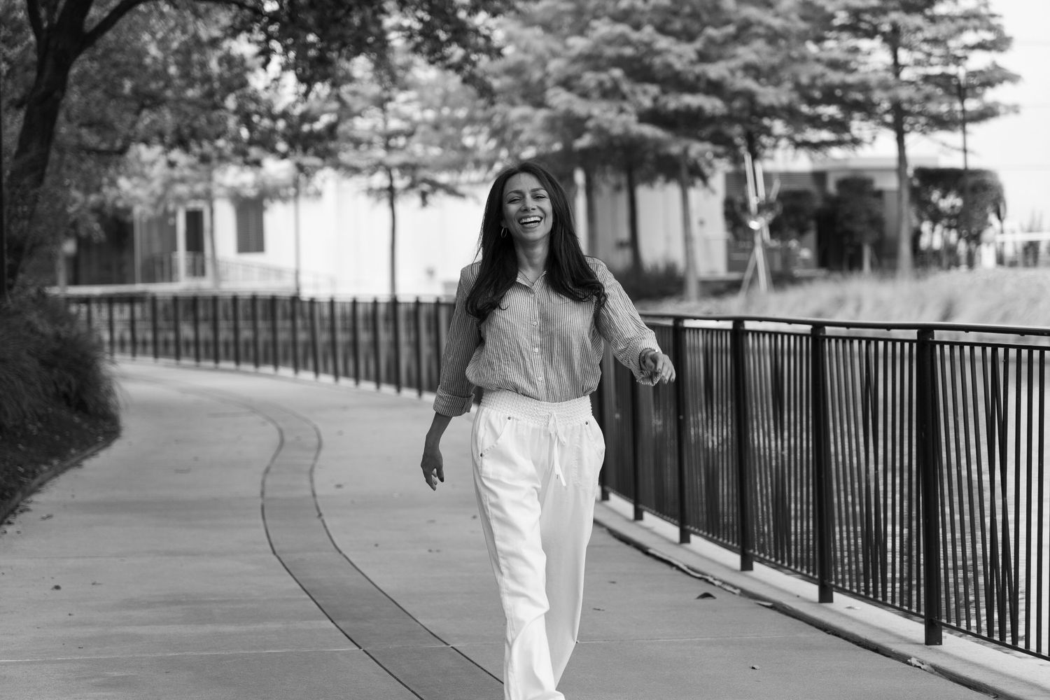 A smiling woman walks along a waterfront path in white pants and a striped top in a black and white photo.