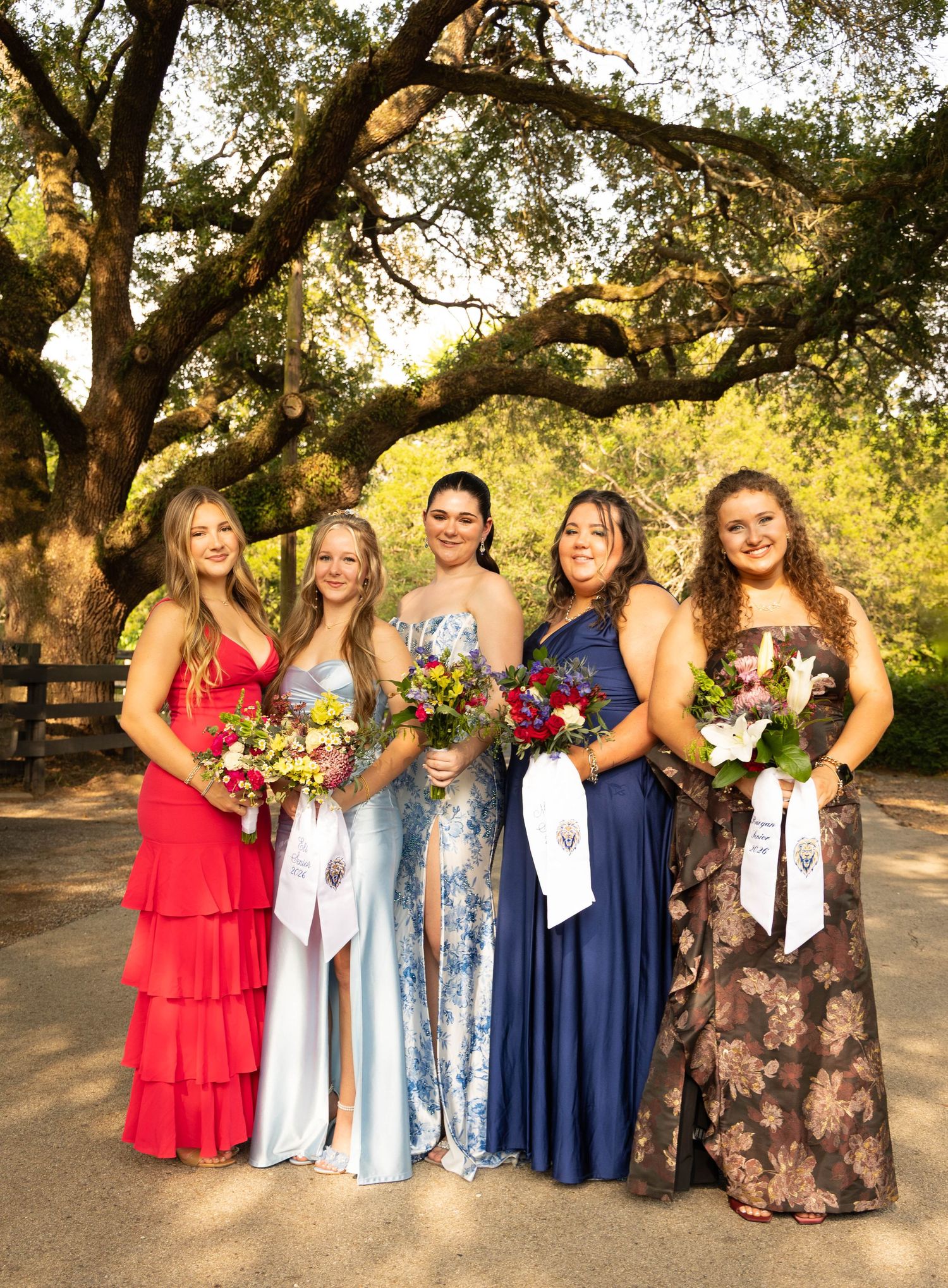 Five young women in formal gowns holding bouquets posing together outdoors under large oak tree.