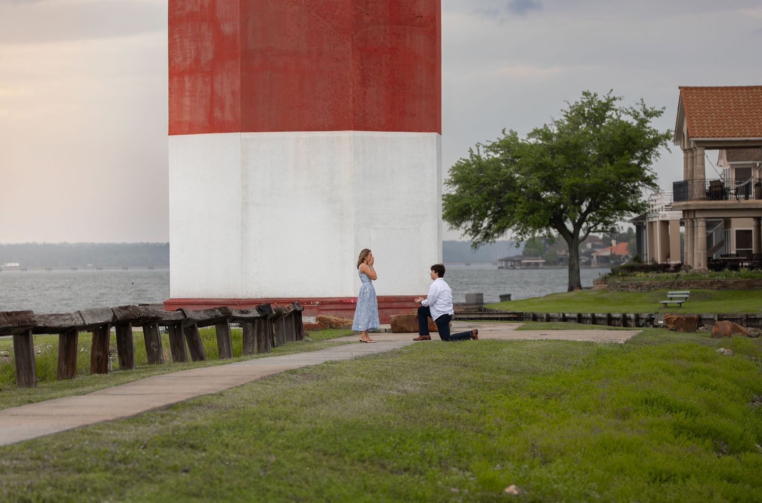 Man proposes on one knee to woman beside a red and white lighthouse near the ocean at sunset.