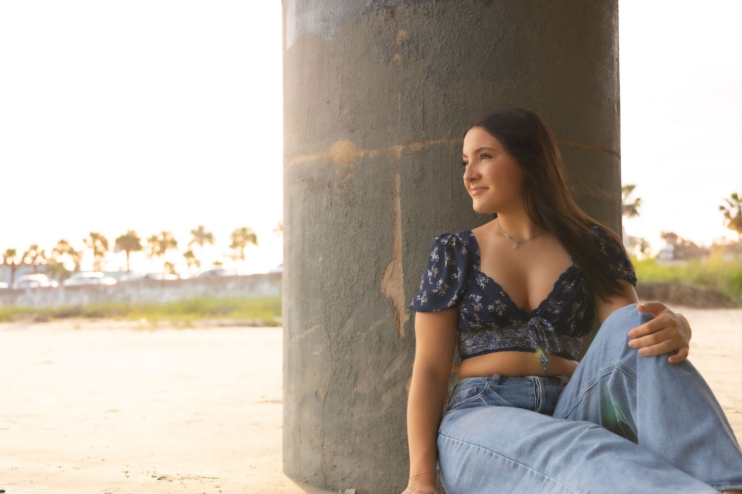 Young woman in floral crop top and jeans sitting against concrete pillar on sandy beach at sunset.