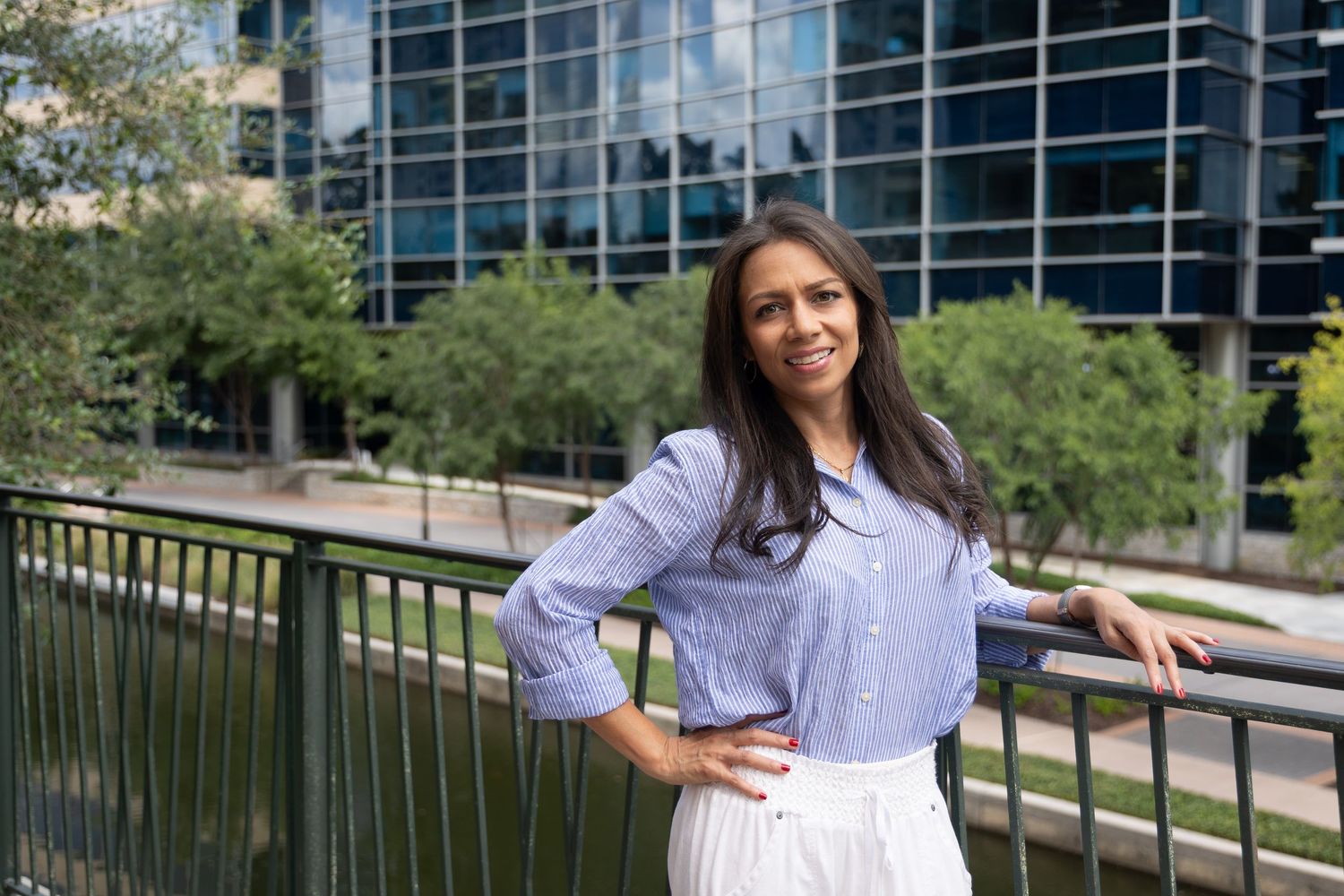 Young woman in blue striped shirt and white skirt posing on urban bridge with modern glass buildings and trees behind her.